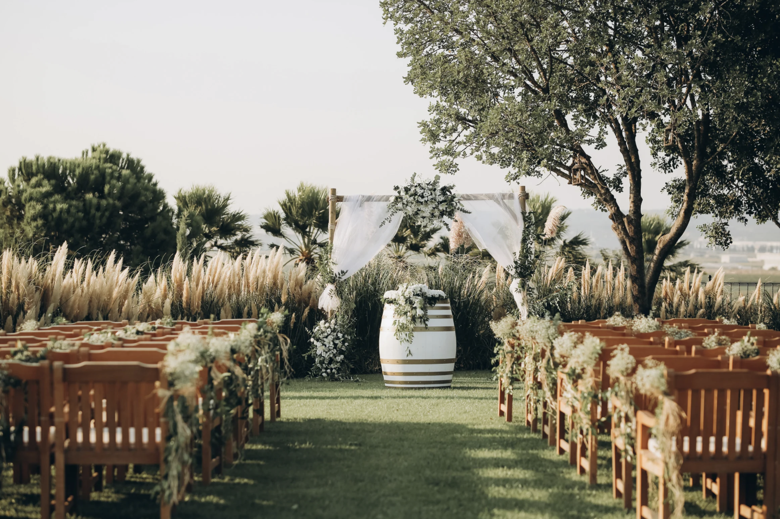 Outdoor wedding ceremony setup with wooden chairs decorated with flowers, a floral arch, a barrel with flowers, and a tree in the background.