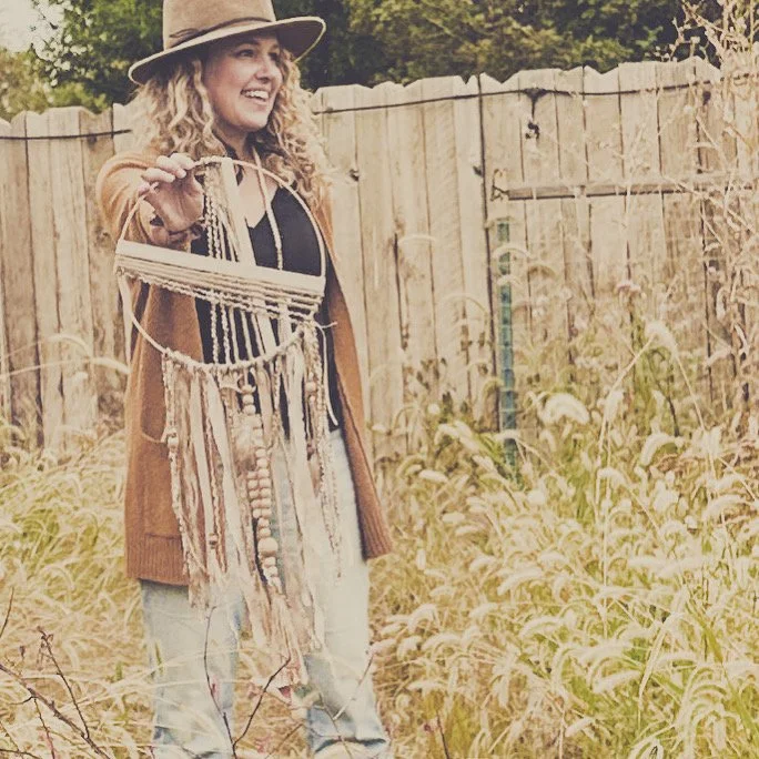 A woman with curly hair wearing a hat and cardigan, smiling while holding a dreamcatcher in a garden with a wooden fence and plants.