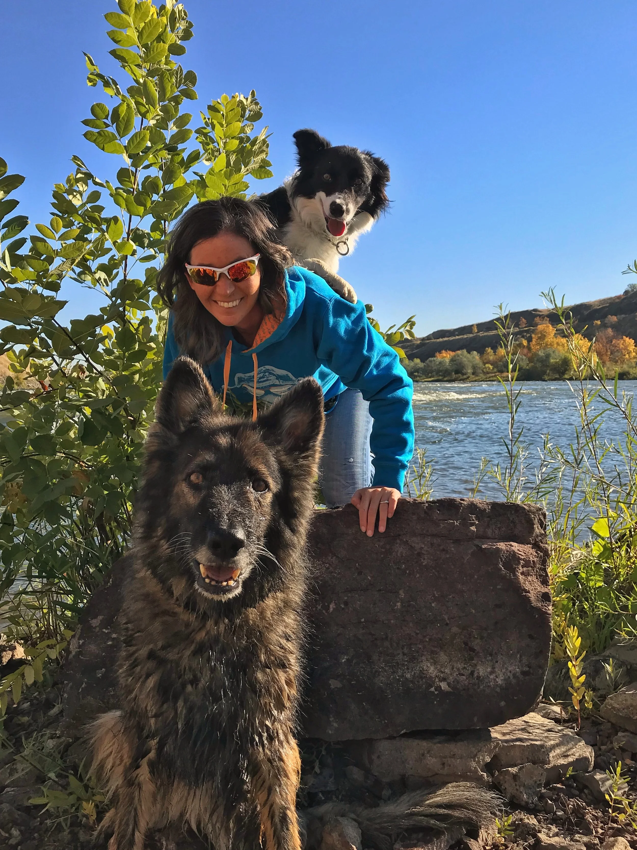 A woman with dark hair, wearing sunglasses and a blue hoodie, is smiling while outdoors near a river. She has two dogs with her: a fluffy, brown and black dog in front and a black-and-white dog on her back. The background shows green bushes, a river,