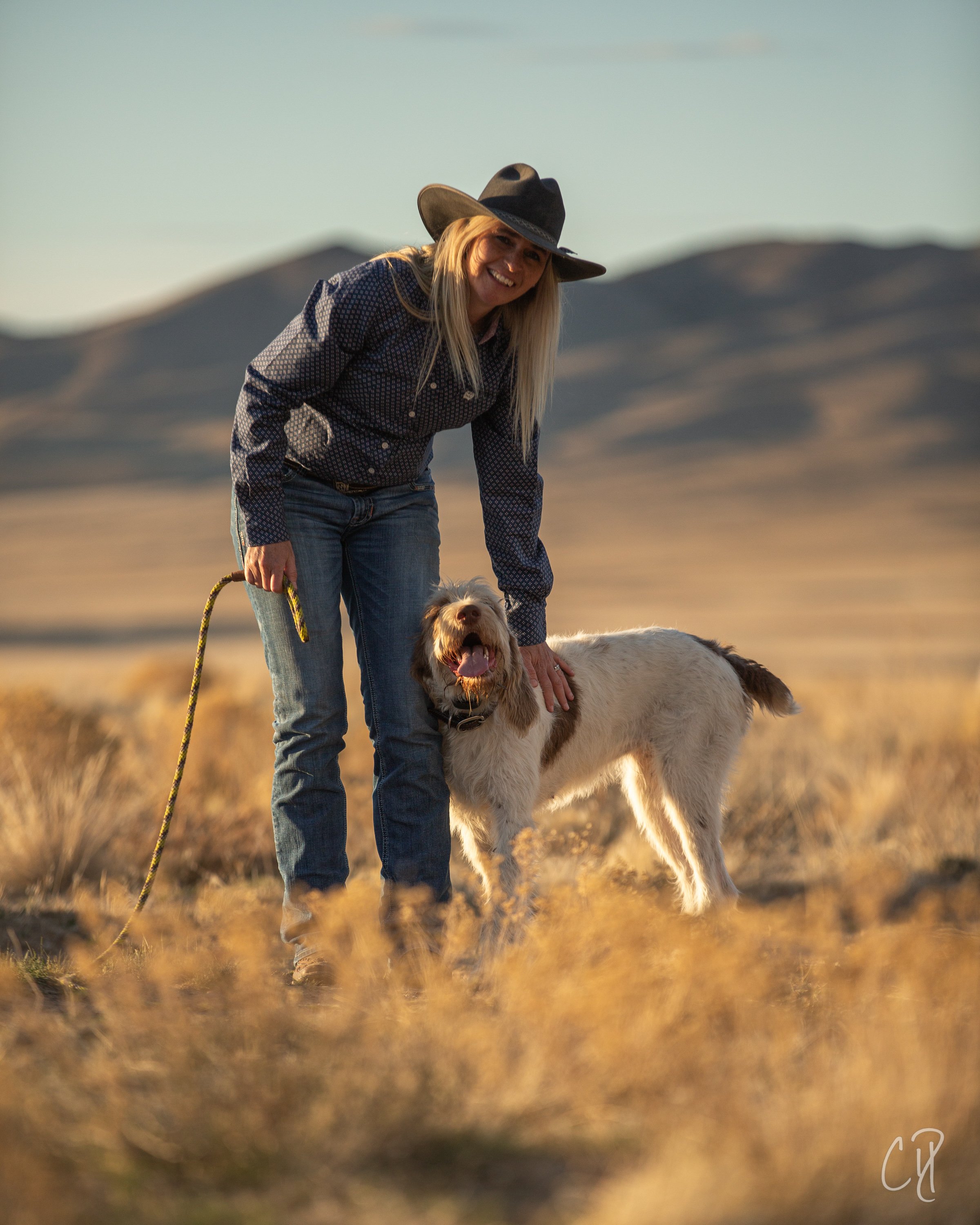 A woman wearing a black cowboy hat, a checkered shirt, and jeans stands outside in a dry, grassy landscape with mountains in the background. She is smiling and bending down to pet a large, fluffy dog that is standing on all fours, looking happy with 