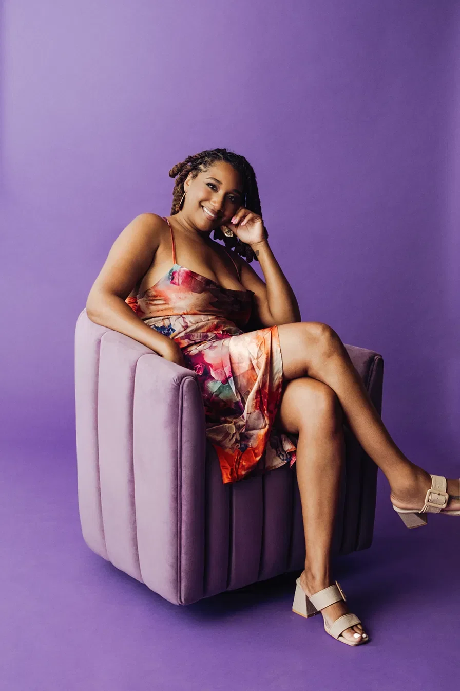 A woman with styled dreadlocks adorned with purple flowers, wearing a black dress and purple tulle skirt, seated on a wooden chair against a beige wall.