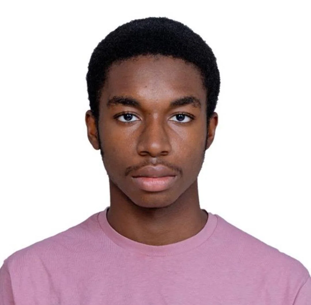 Portrait of a young man with short black hair, wearing a pink T-shirt, looking directly at the camera against a plain white background.