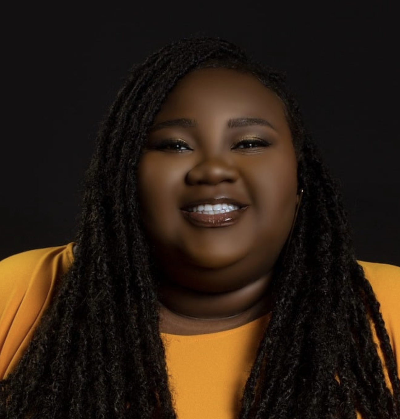Portrait of a plus-sized woman with dark skin and long, textured black hair, smiling, wearing a yellow top against a black background.
