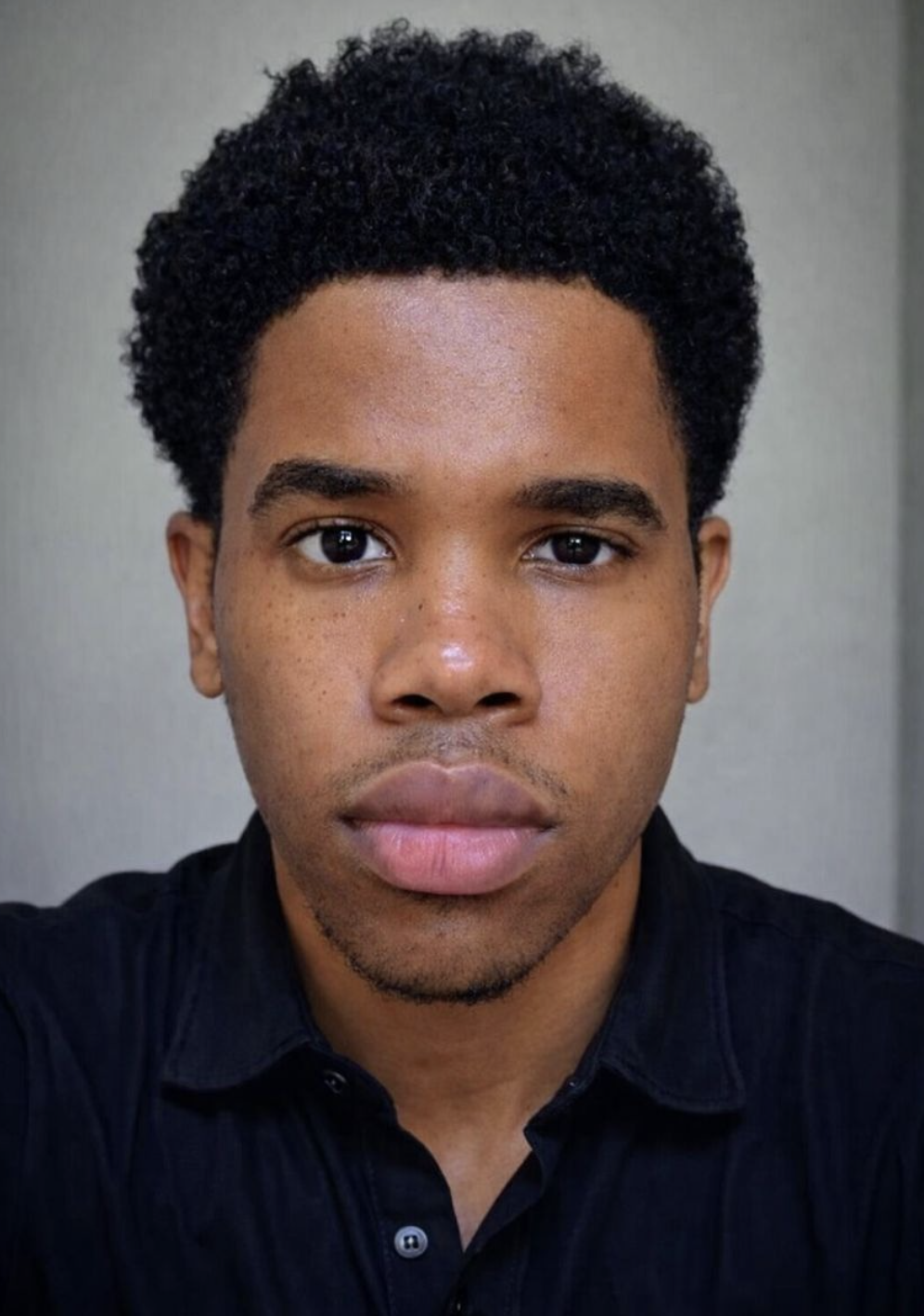 Close-up portrait of a young man with short, curly black hair, wearing a black collared shirt, looking directly at the camera with a neutral expression.