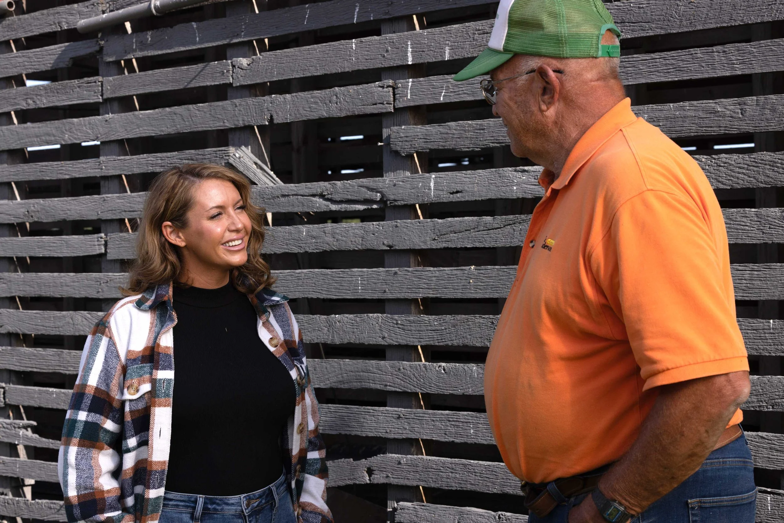 Wezlynn Davis and an older man stand outdoors in front of a weathered wooden fence, smiling and engaged in conversation.