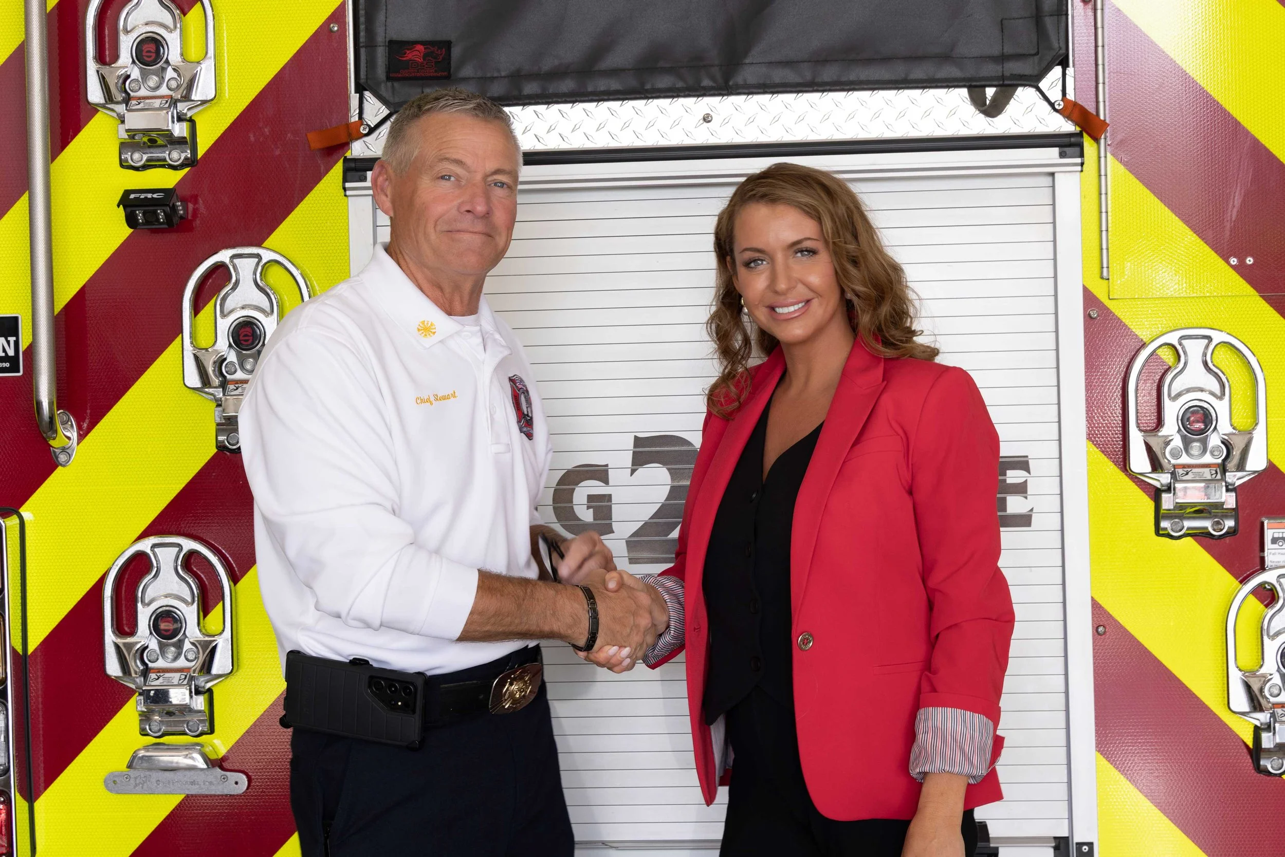 A firefighter in uniform shaking hands with Wezlynn Davis in a red blazer in front of a fire truck.