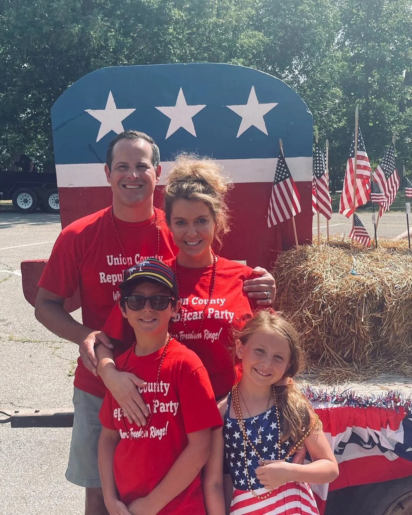 A family of four celebrating Independence Day outdoors, wearing red, white, and blue clothing, standing in front of a float decorated with American flags, hay bales, and patriotic decor, with an American flag-themed backdrop.
