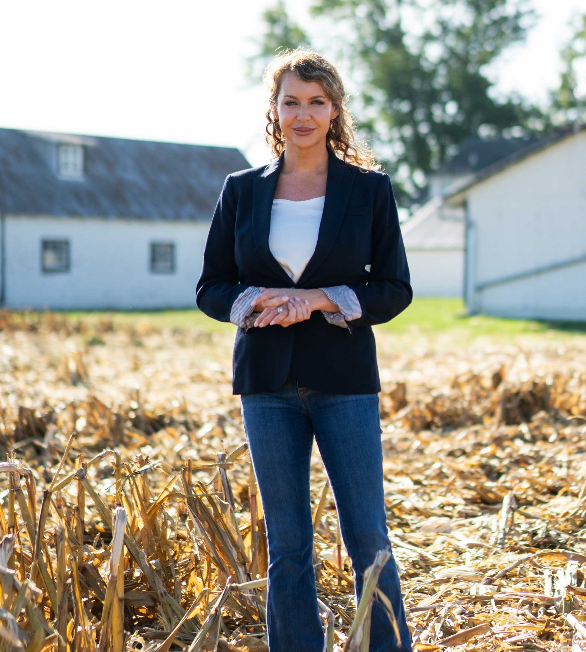 A woman with curly light brown hair, wearing a navy blazer over a white shirt and blue jeans, standing outdoors in a cornfield with dried corn stalks during fall, with trees and farm buildings in the background.
