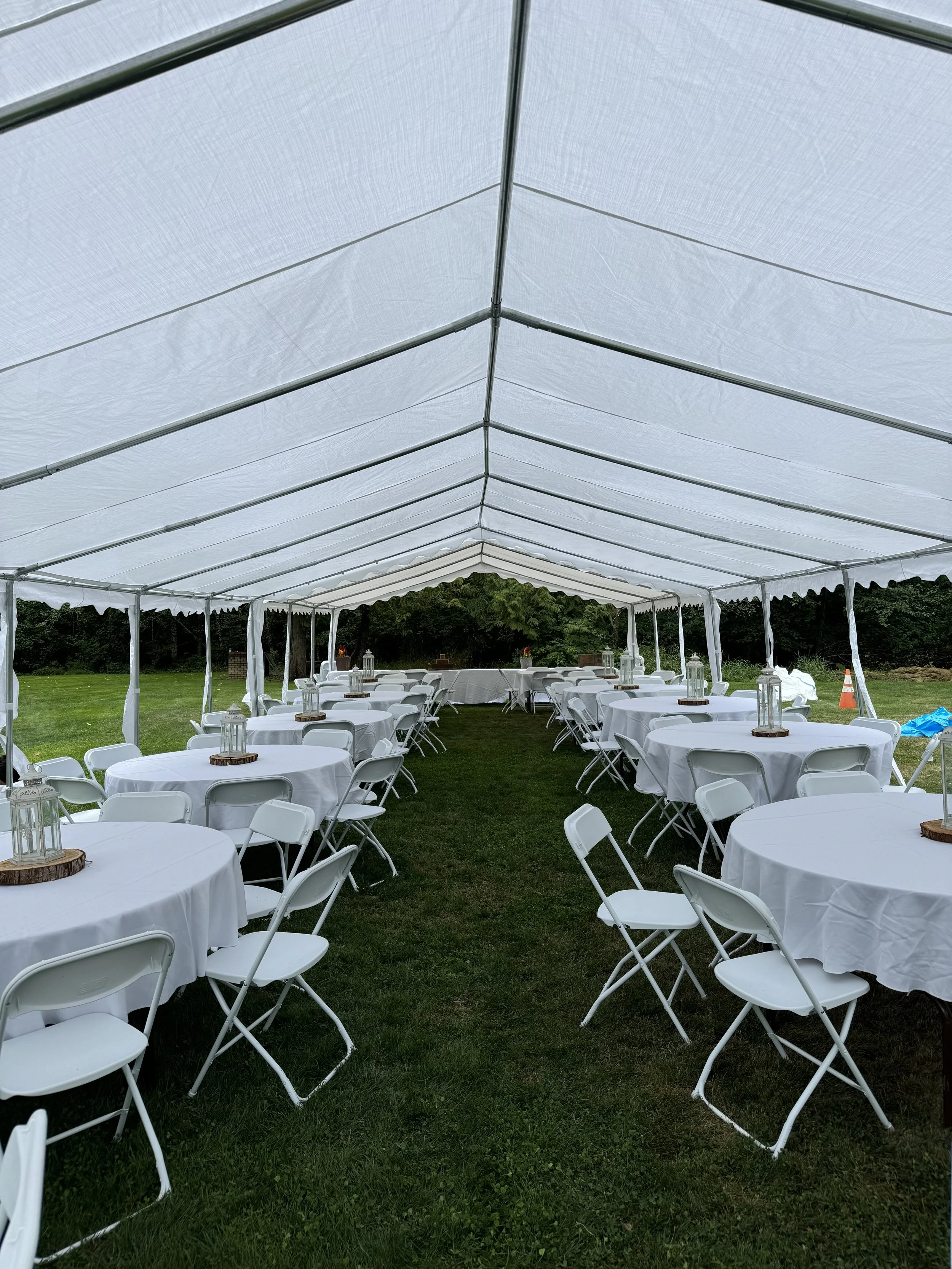 White round tables with white chairs under a outdoor tent, decorated with lanterns on wooden bases, set up for an event on a grassy area.