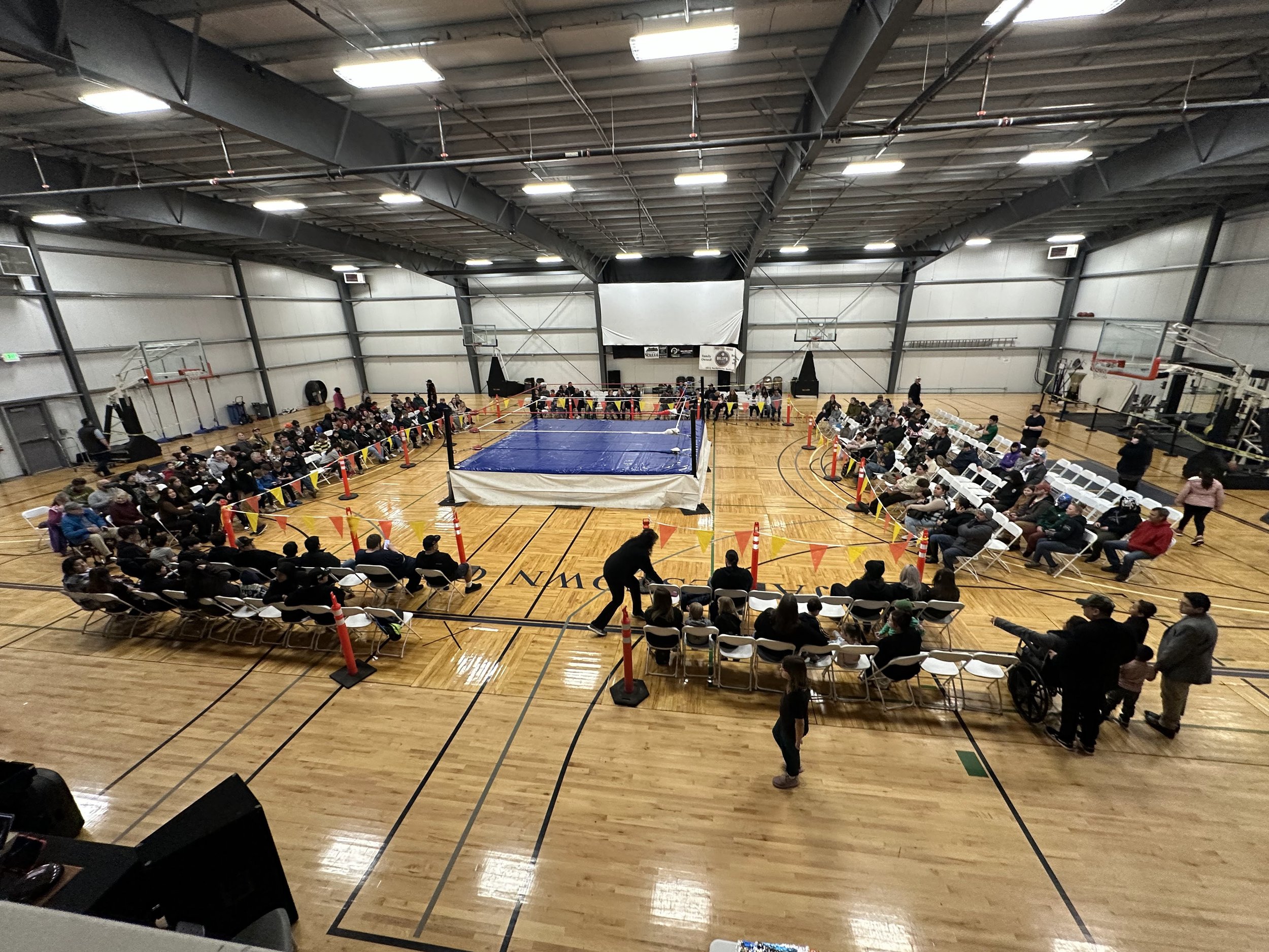An indoor wrestling gymnasium with a wrestling ring at the center, surrounded by rows of chairs filled with spectators. The gym has a high ceiling with metal beams and doors on the sides.