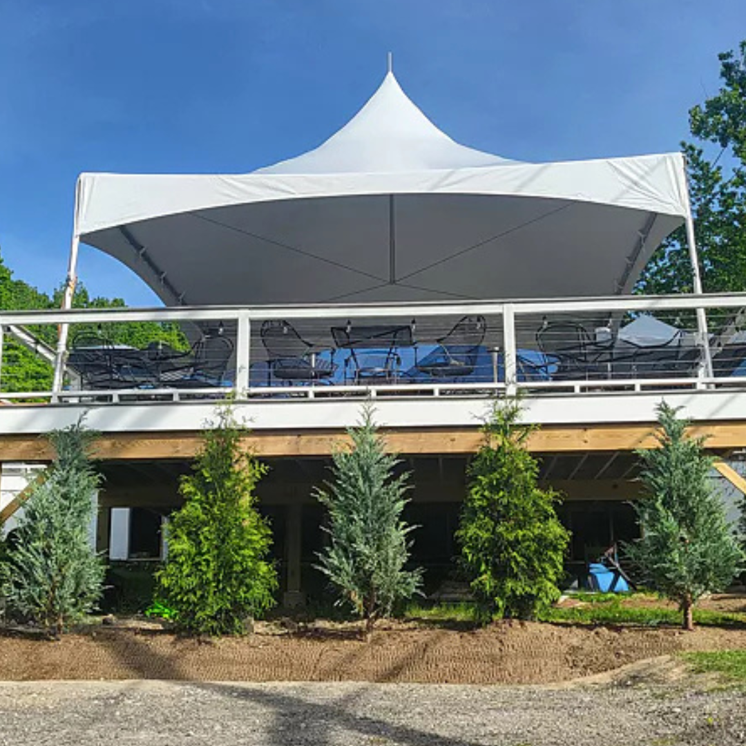 A large white outdoor canopy tent with high peak, covering a balcony with tables and chairs, situated above a row of green trees and shrubs on a sunny day.