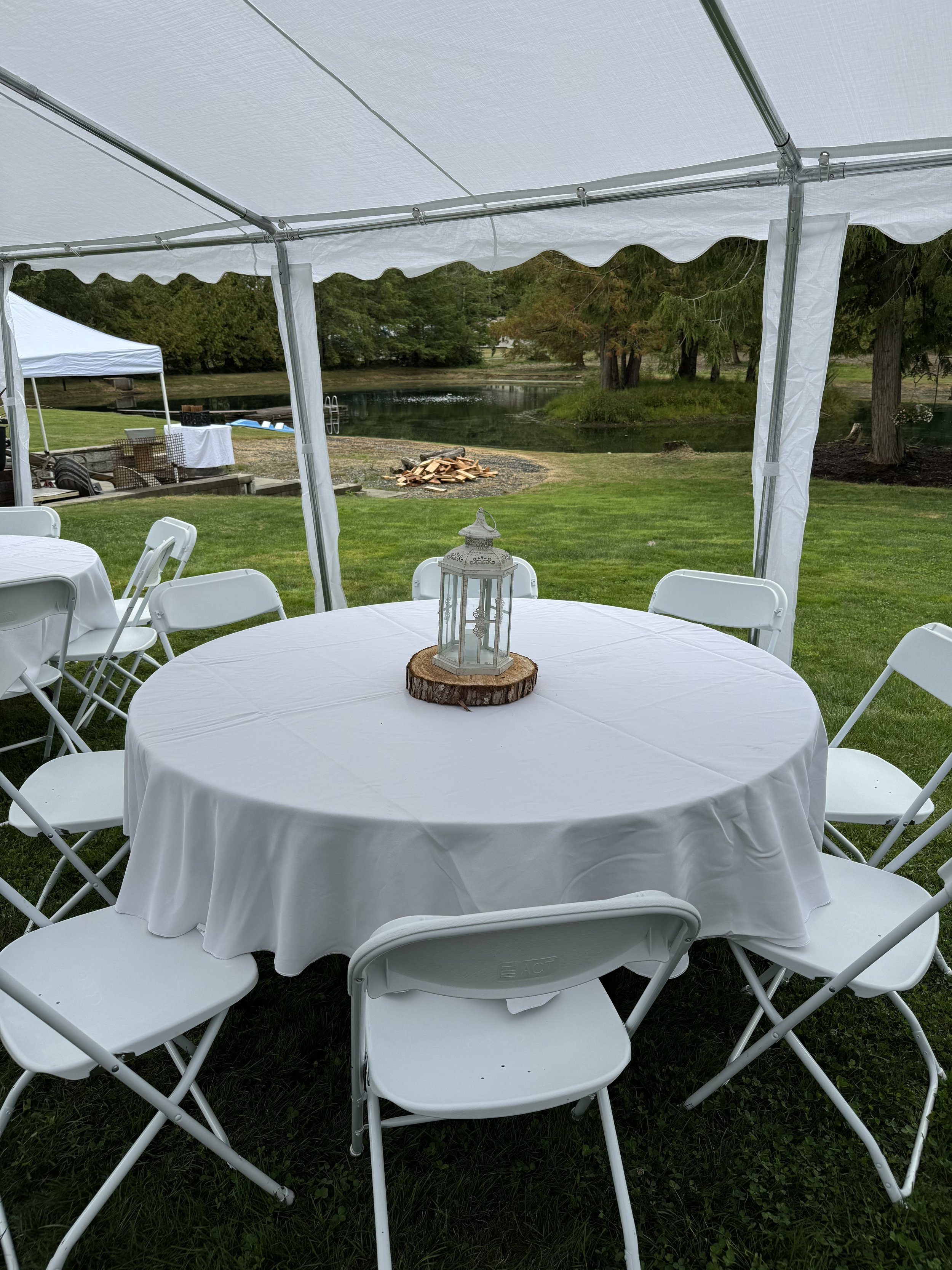 Outdoor event setup with a round table covered in a white tablecloth, white folding chairs, and a lantern centerpiece on a wooden slab. There is a white tent canopy and a scenic view of a pond, trees, and a firepit in the background.