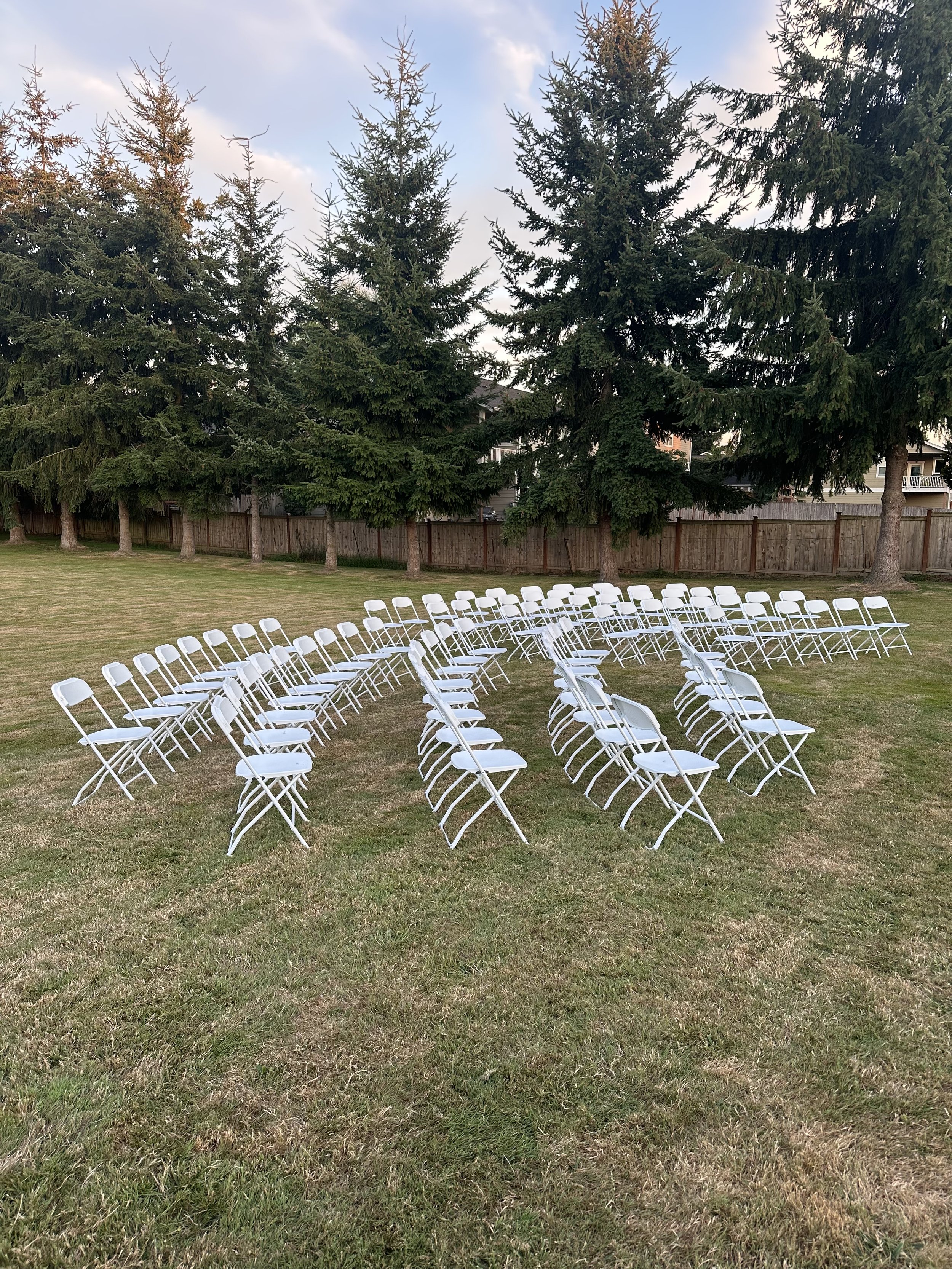 Rows of white folding chairs arranged on a grassy lawn with pine trees and a wooden fence in the background, preparing for an outdoor event.
