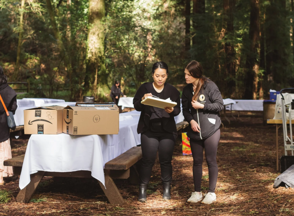 Two women standing outdoors in a wooded area, reviewing documents over a table with Amazon boxes, surrounded by tables covered with white cloths.