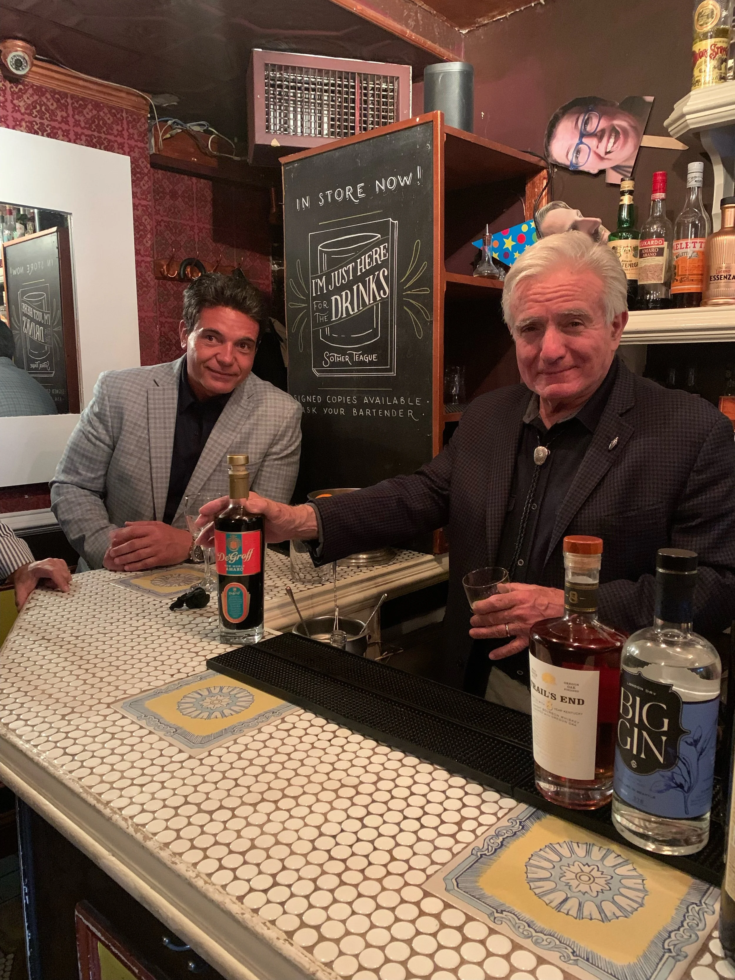 Two men sitting at a bar, one with gray hair and the other with dark hair, shaking hands and smiling. The bar has a white tiled countertop with drink bottles and glasses. There is a chalkboard sign behind them that says 'I'm just here for the drinks' and 'In store now!'. The background includes a mirror, a red patterned wall, and some decor, including a large photo of a woman's face and various bottles of alcohol.