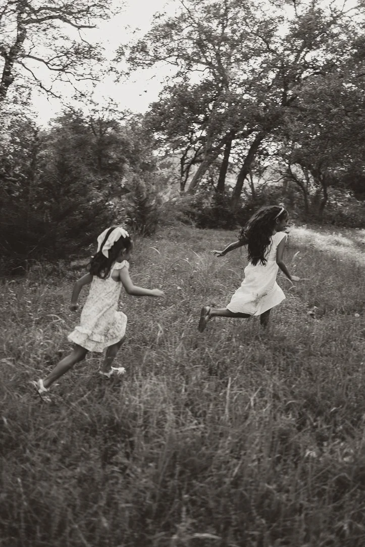 Siblings run through a sun-dappled forest path, dresses flowing behind them, as they chase the last light of the day.
