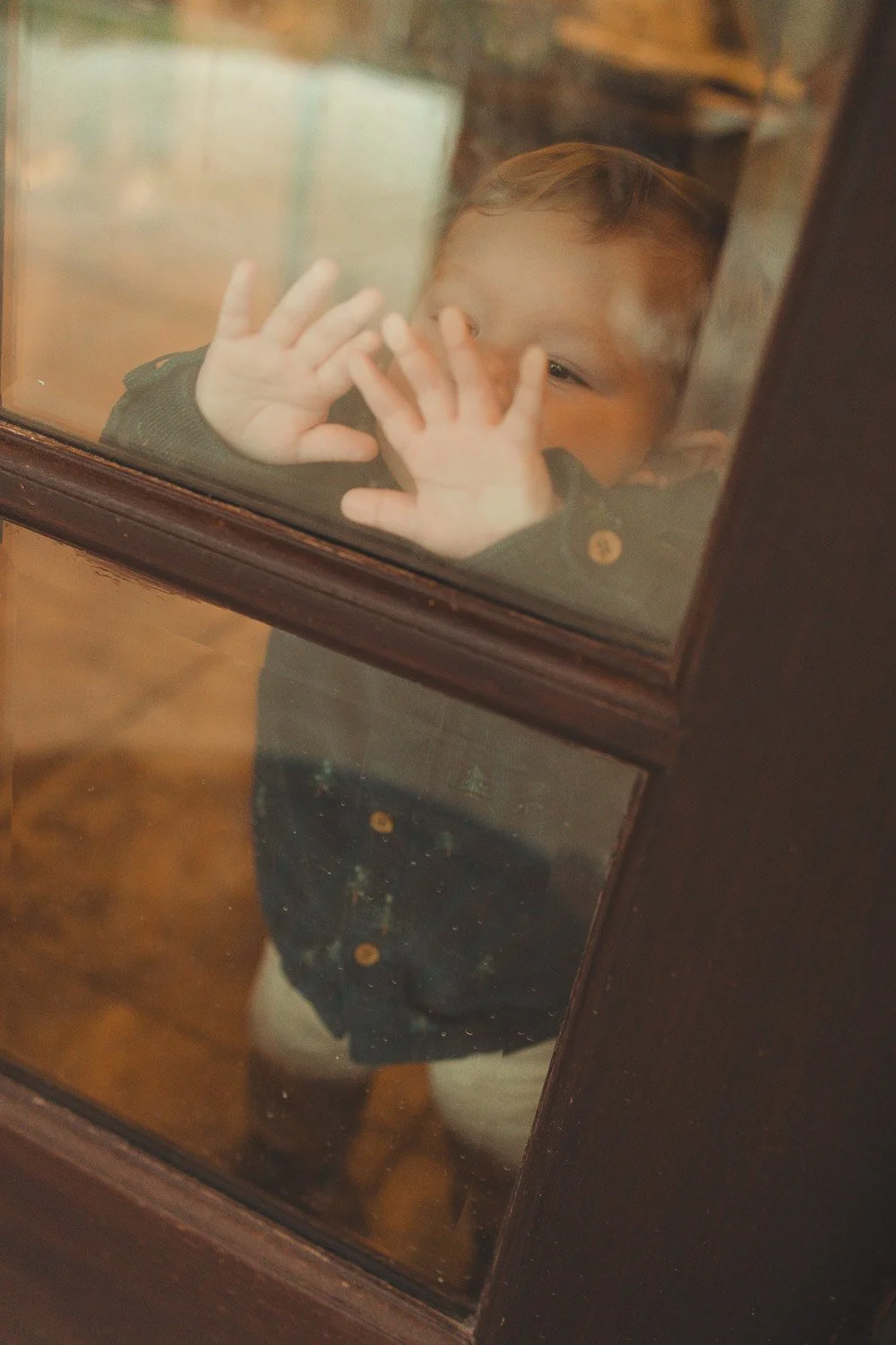 A young child with light brown hair looking through a glass door, pressing their hands and face against the glass and peering outside.