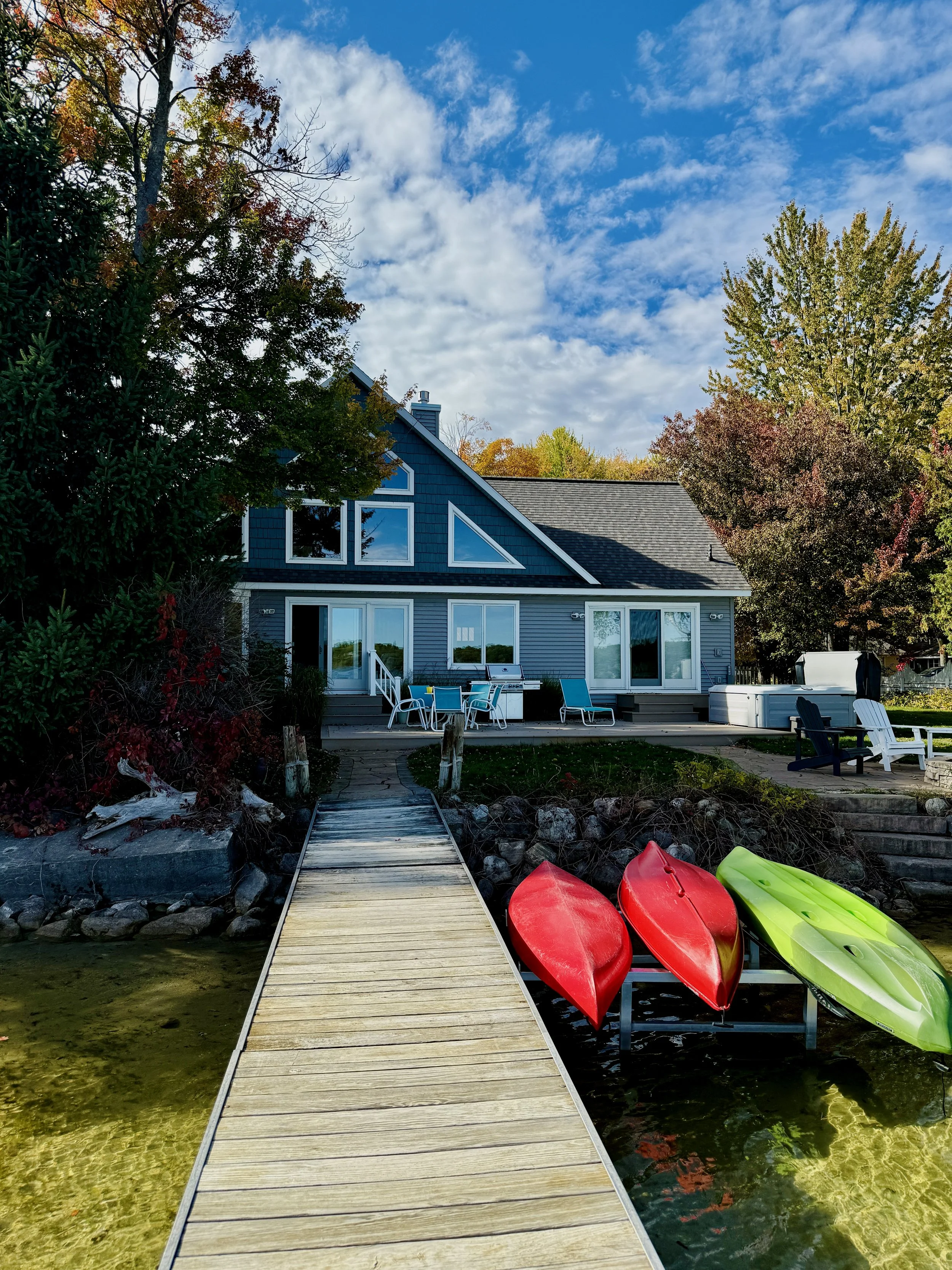 A house with blue siding and large windows, surrounded by trees with fall foliage. A wooden dock extends over a small lake or pond with three kayaks—two red and one green—resting beside it. There is outdoor seating on the deck, including a table with