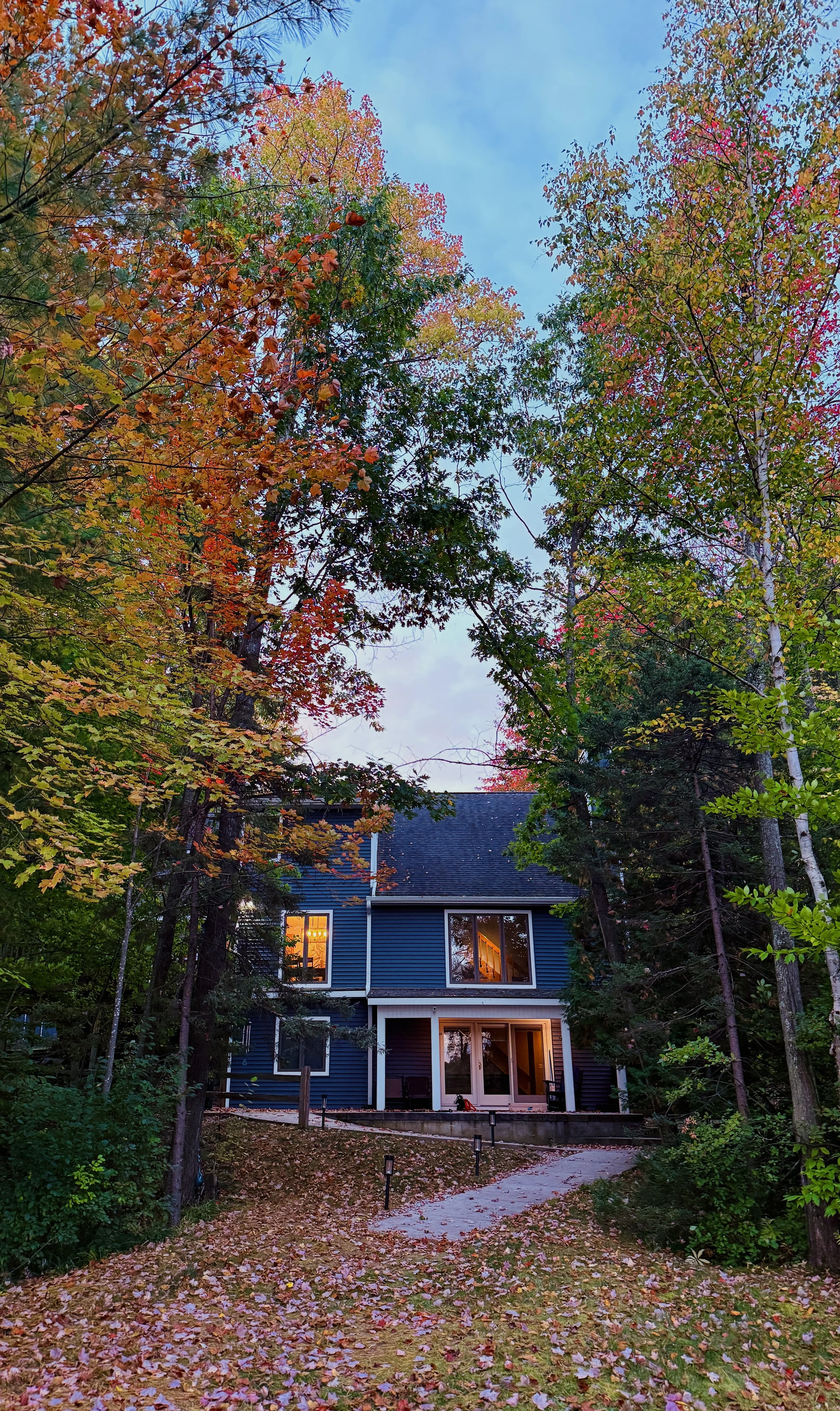 A house with blue siding in a wooded area surrounded by trees with fall foliage, with warm lights visible through the windows, and a pathway leading to the house.