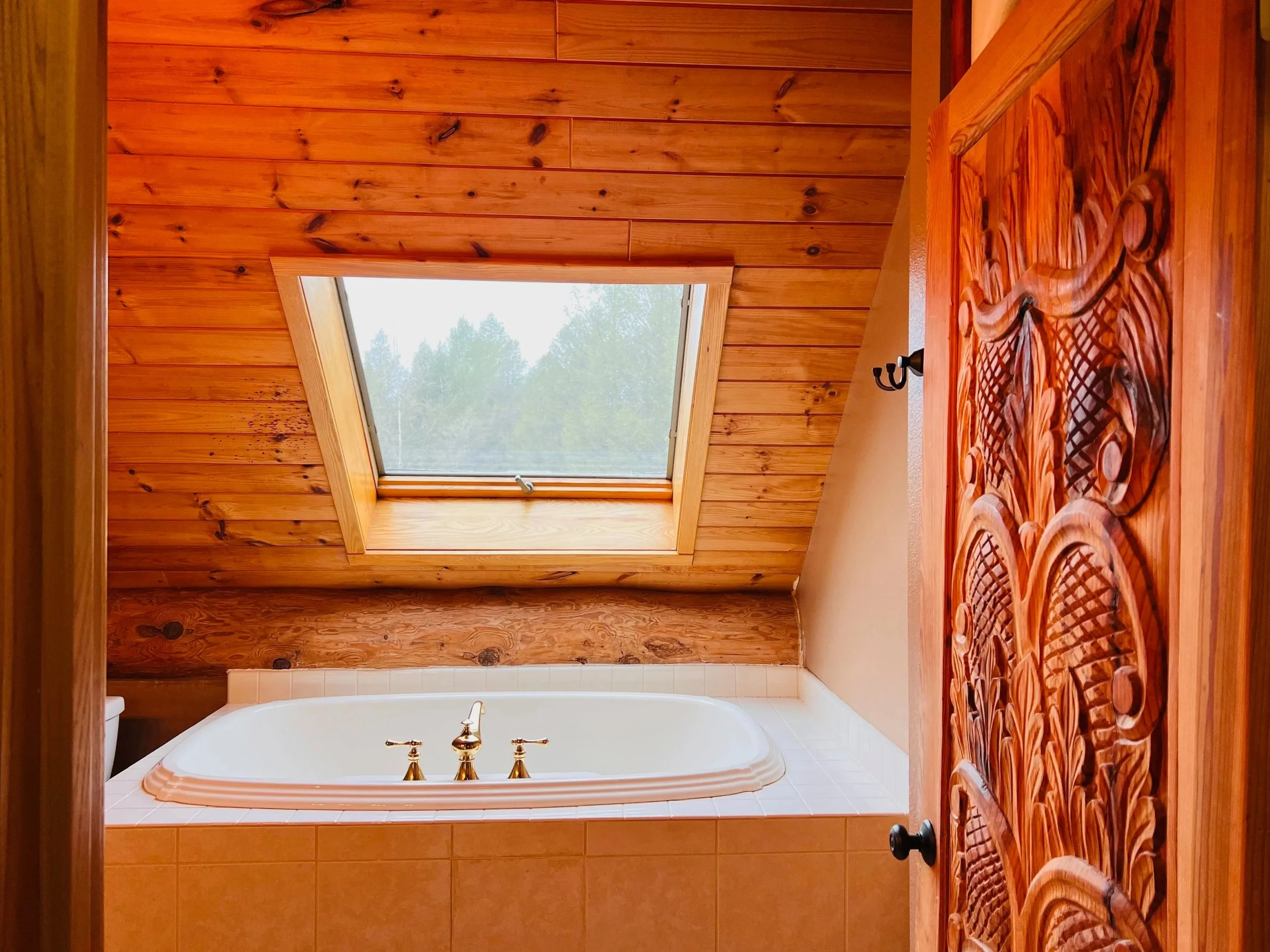 A bathroom with a wooden ceiling and a skylight window, featuring a white bathtub with brass fixtures and a carved wooden door.
