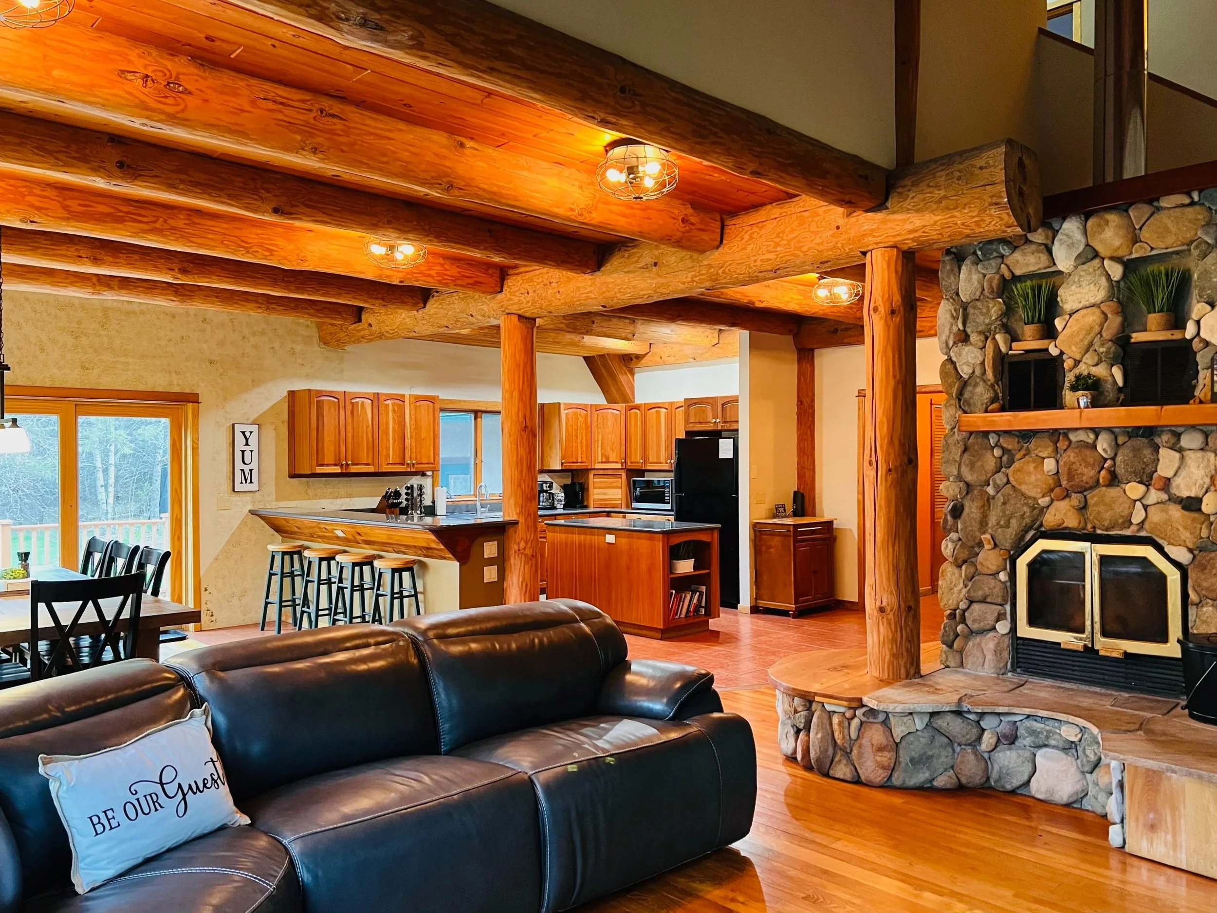 Interior of a cozy living room and kitchen area with wooden ceiling beams, a stone fireplace, leather couch with a decorative pillow that says 'BE OUR GUEST', and sliding glass doors leading outside.