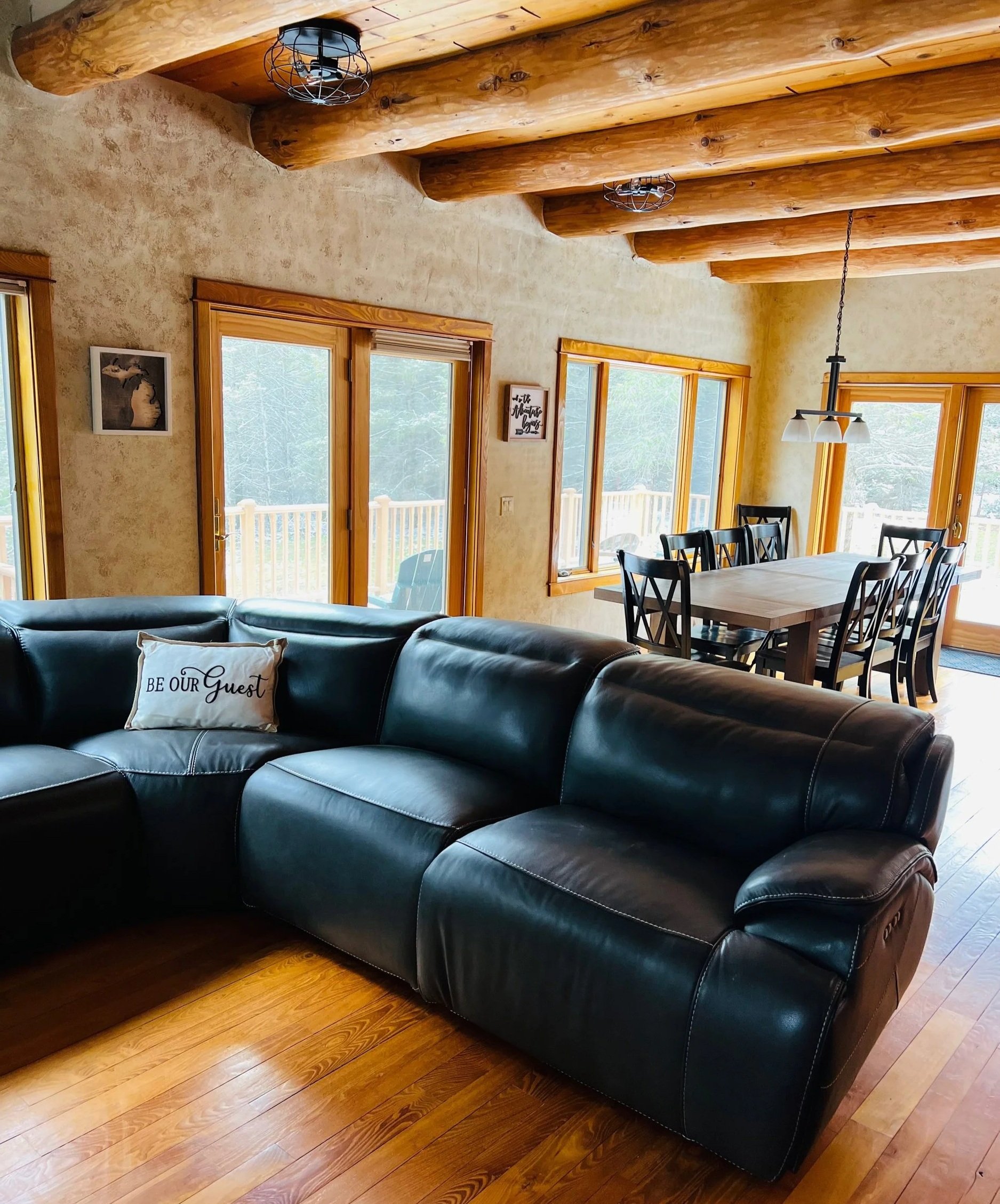 Living room with leather sofa, wooden ceiling beams, and large windows with a view of a deck outside.