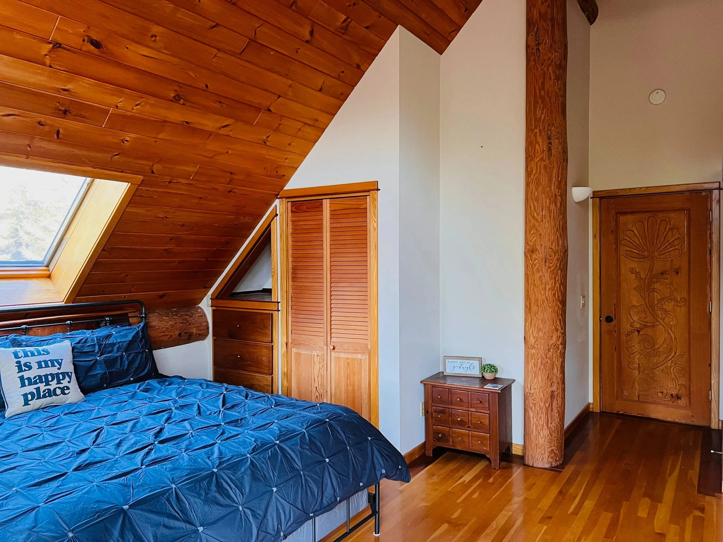 A cozy bedroom with a sloped wooden ceiling and hardwood floors. The bed has a blue quilt and a decorative pillow that says 'this is my happy place.' There is a small wooden nightstand with a sign and a small plant on top. A large skylight window is 