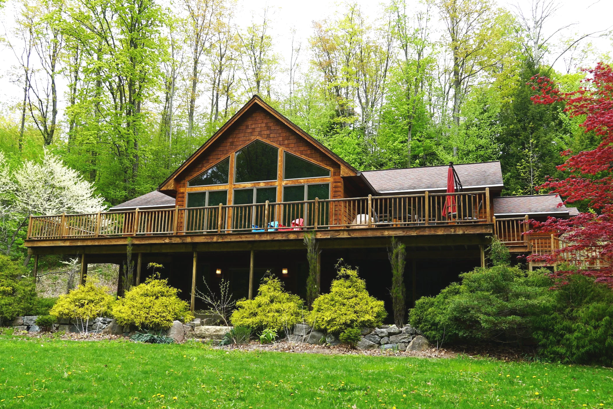 Wooden house in a green forest with a large deck and outdoor furniture.