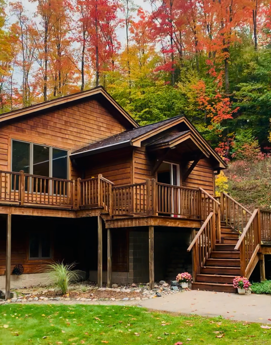 A wooden house with a deck, stairs, and a lush green lawn in front, surrounded by colorful autumn trees with red, orange, and yellow leaves.