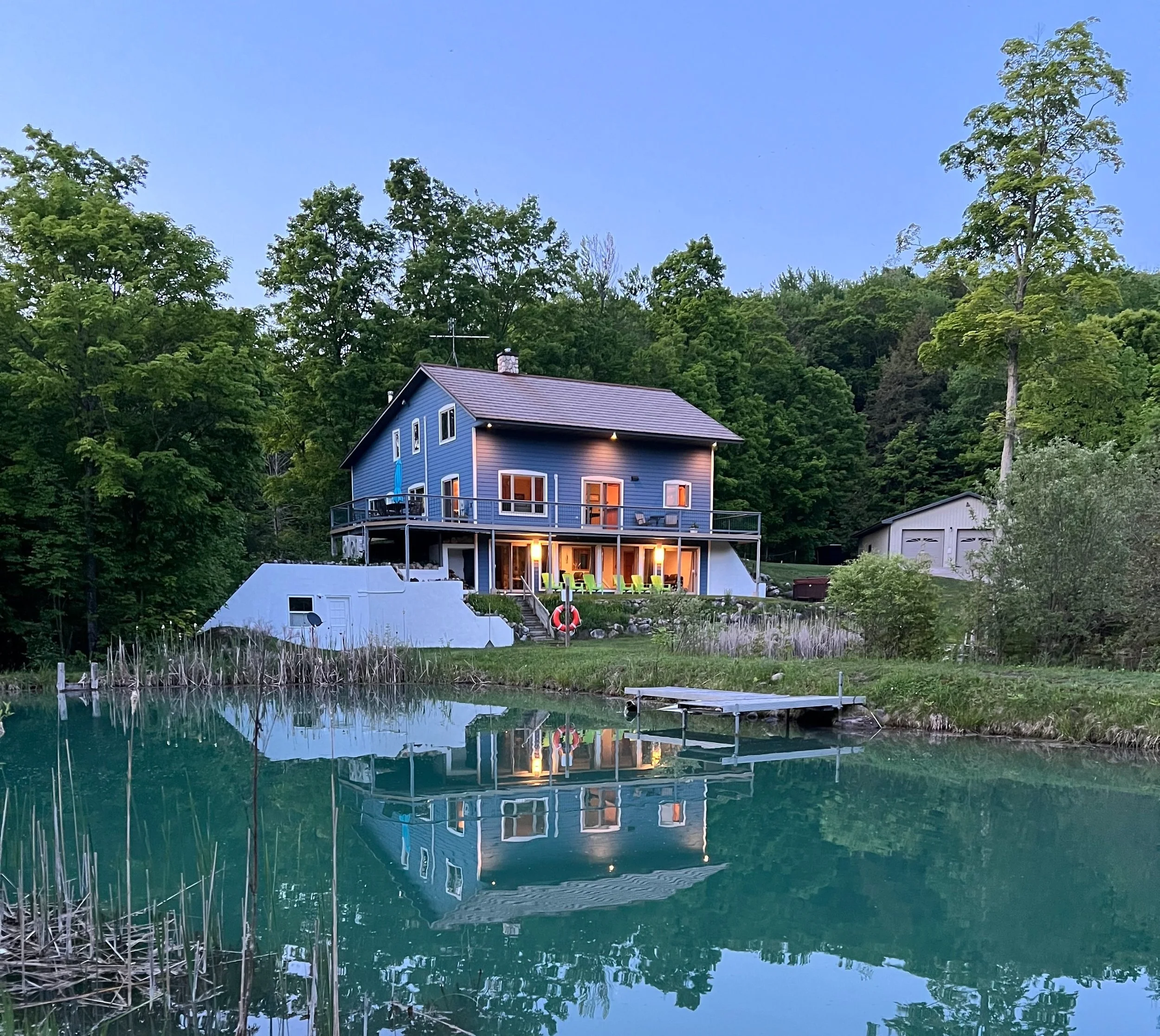A blue house with a deck overlooking a pond, surrounded by green trees at dusk.
