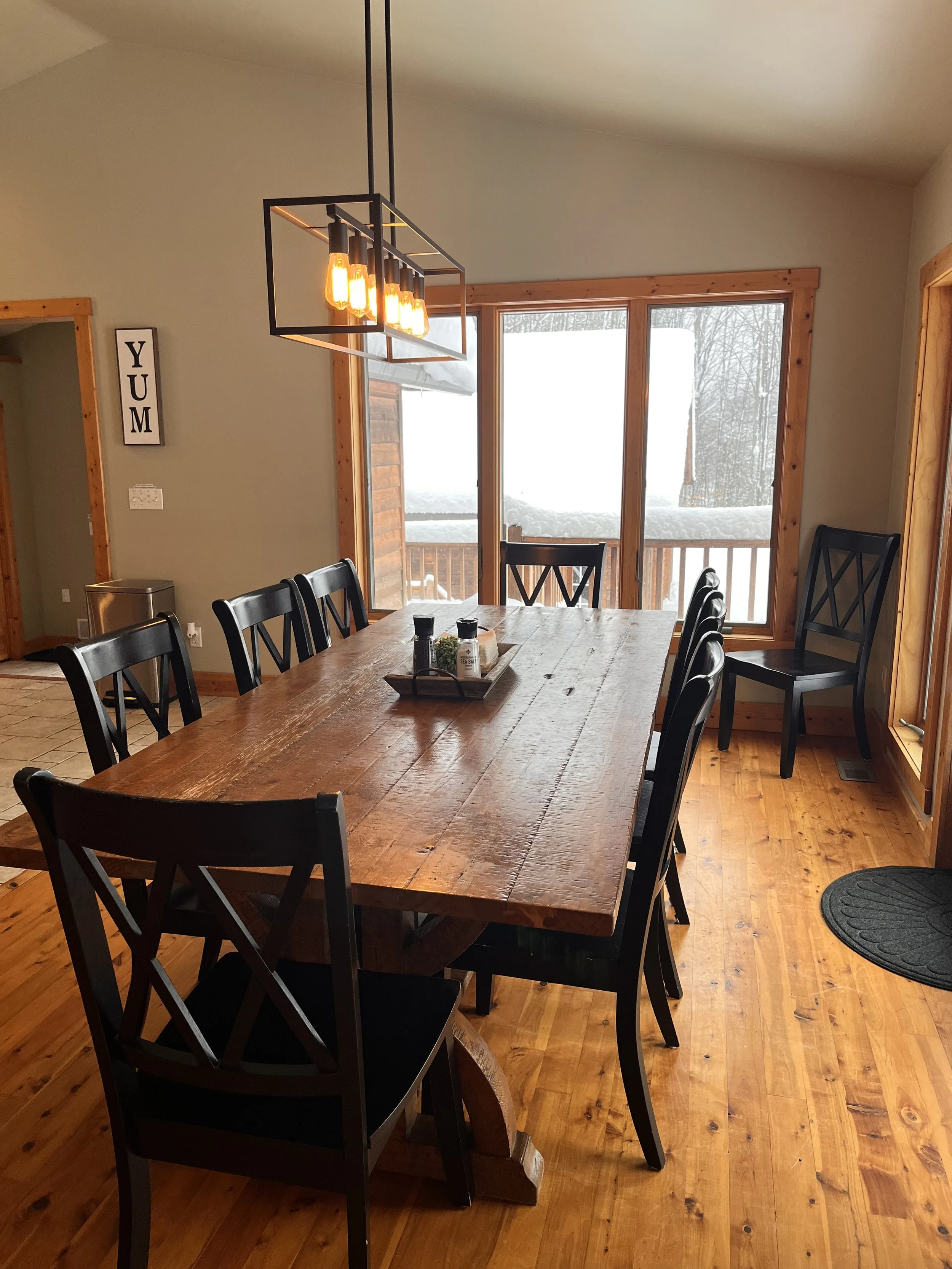 A rustic dining room with a large wooden table, black chairs, and a modern rectangular chandelier. Large windows show snow outside, and there is a wall sign that says 'YUM'.