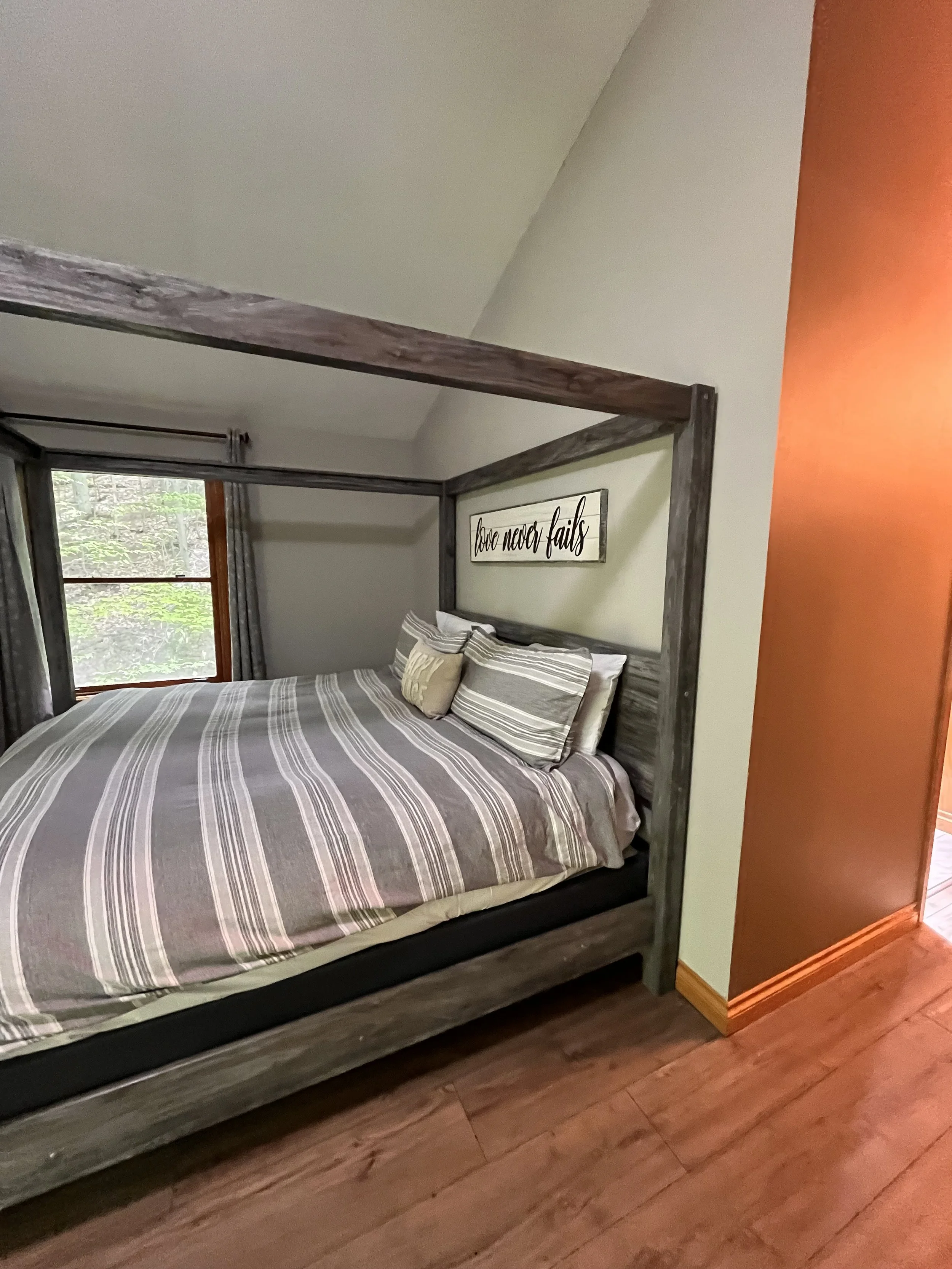 A cozy bedroom featuring a four-poster bed with a wooden frame, striped bedding, multiple pillows, and a wall sign that reads 'love never fails.' A window with curtains is visible, providing natural light.