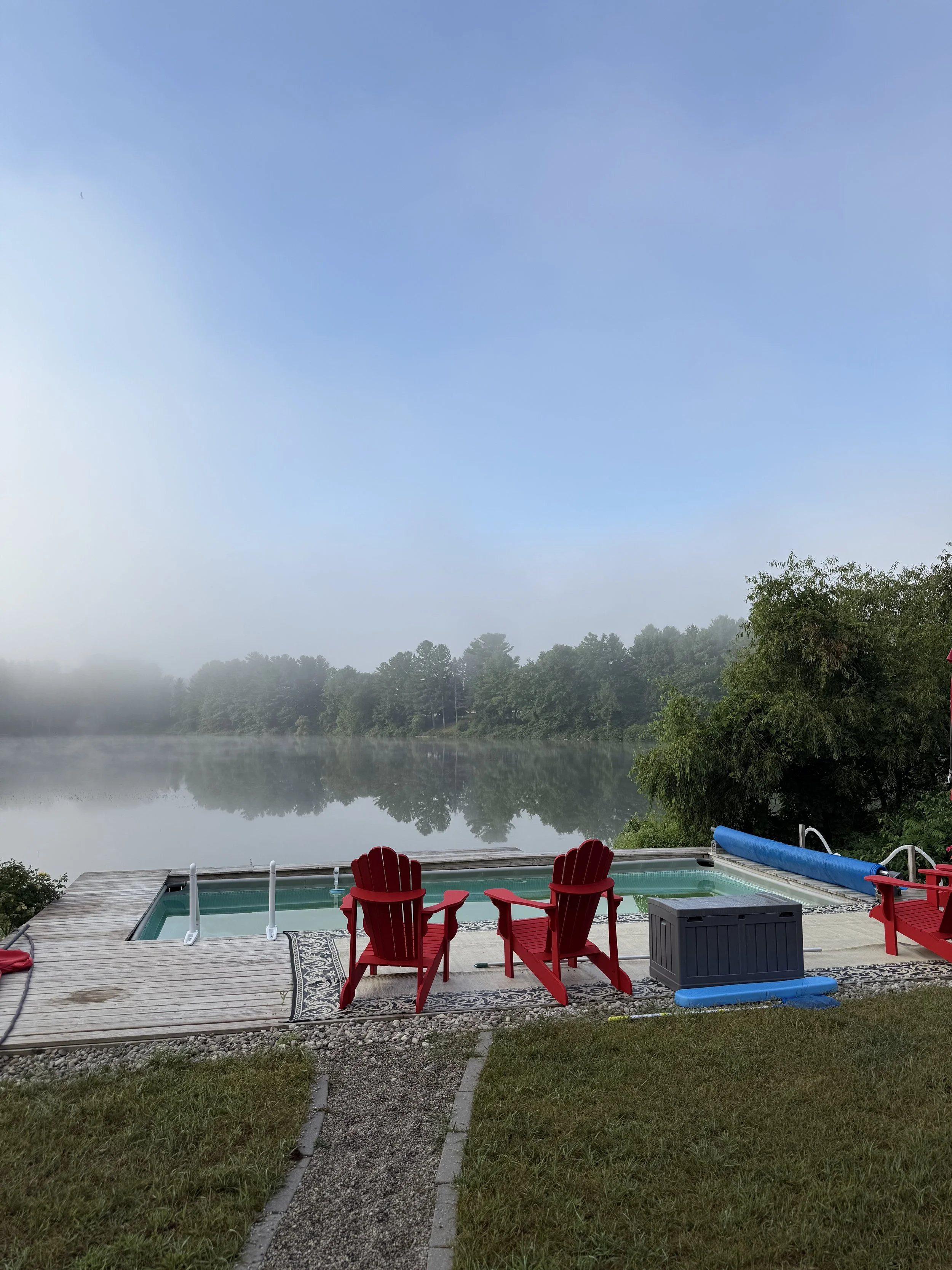 A lakeside view with a small hot tub, two red Adirondack chairs, and a view of foggy water and trees in the background on a clear day.