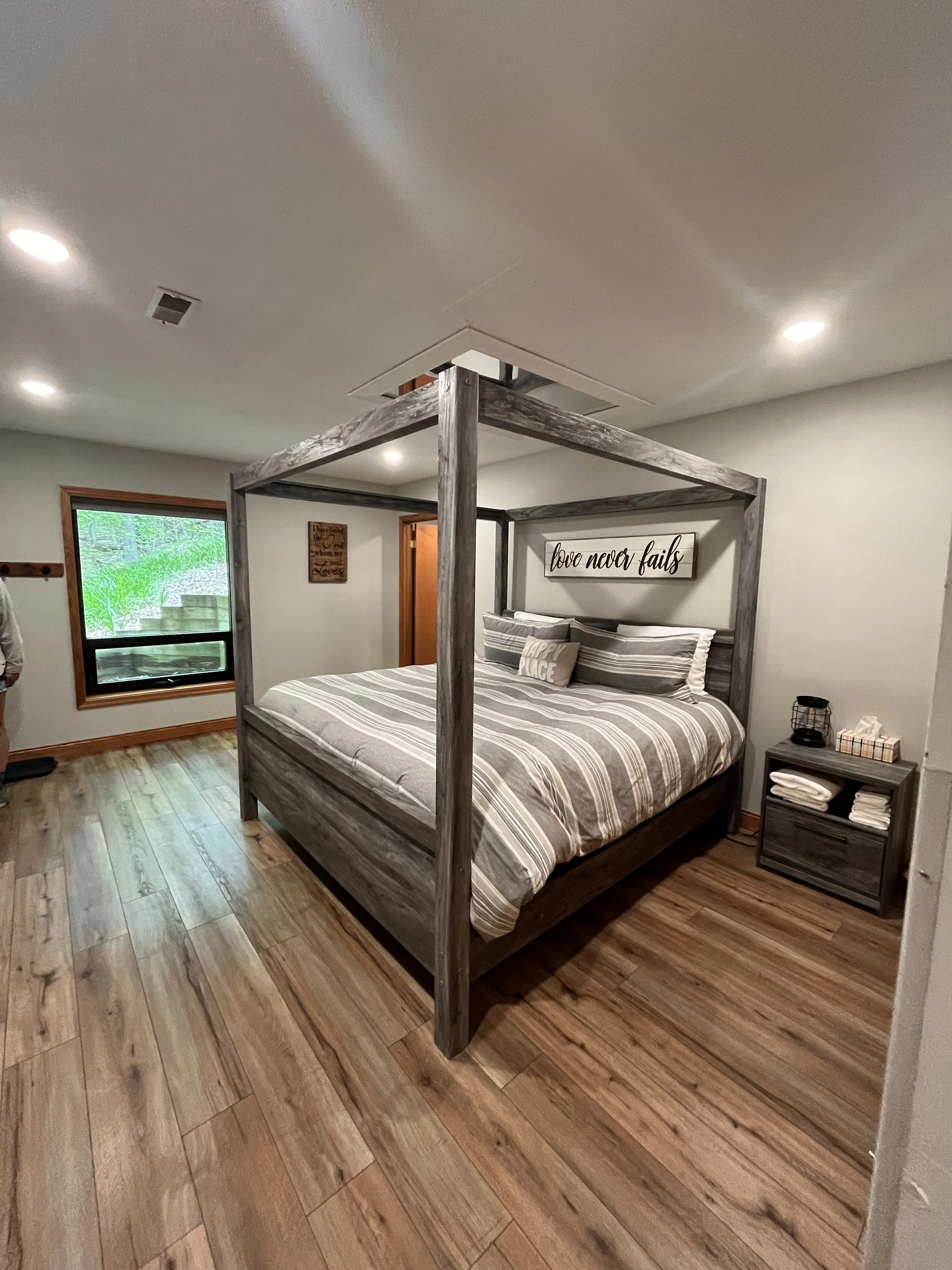 Bedroom with a four-poster rustic wood bed, striped bedding, and decorative pillows with a picture frame that says "love never fails" on the wall. Has a wooden nightstand with towels and a lamp. Window showing greenery outside, wooden floor, and ceil