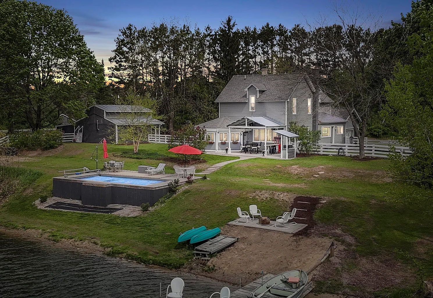 A backyard scene featuring a house with a large deck, a hot tub, a swimming pool, a patio with outdoor furniture and a red umbrella, a sandy beach area with boats and chairs, and trees surrounding the yard.