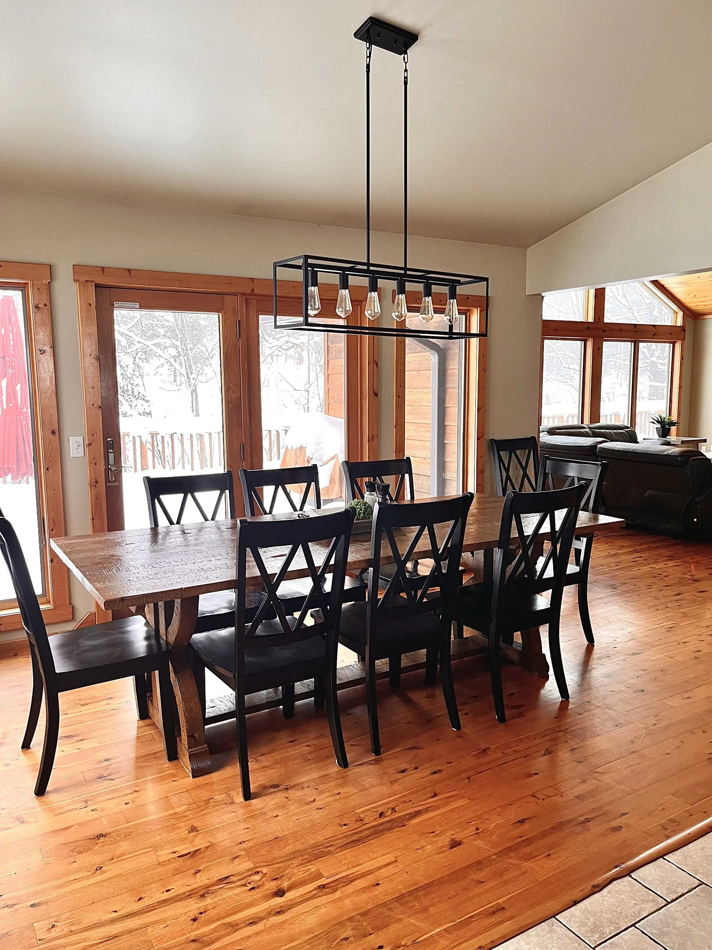 A dining room with a large wooden table and eight black chairs, wooden floors, a modern rectangular chandelier, and sliding glass doors leading to a snowy outdoor area.