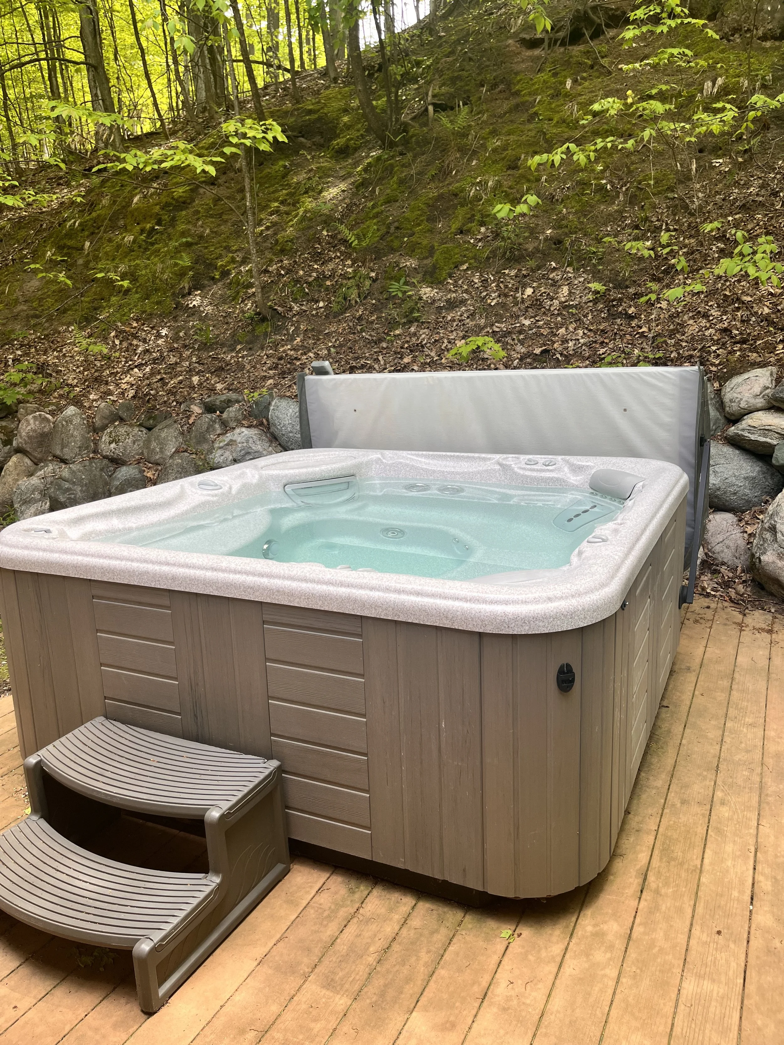 An outdoor hot tub with water, set on a wooden deck, surrounded by rocks and trees in a forested area.