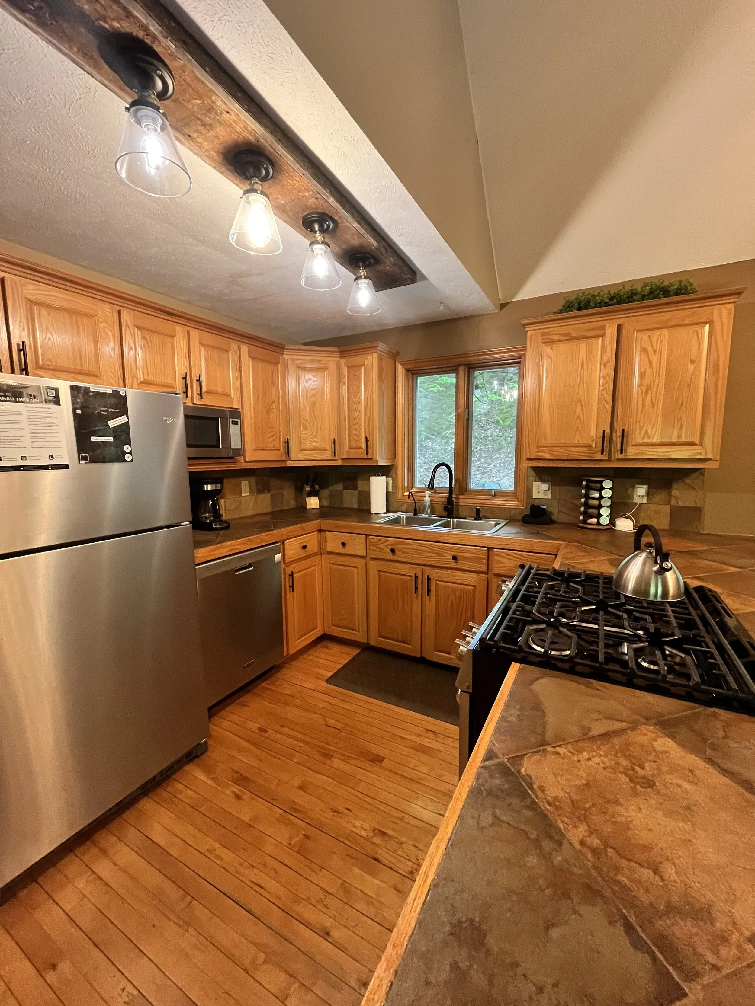 Kitchen with wooden cabinets, stainless steel refrigerator, microwave, dishwasher, black stovetop, and a window above the sink. Wooden flooring and a chandelier with four light bulbs.