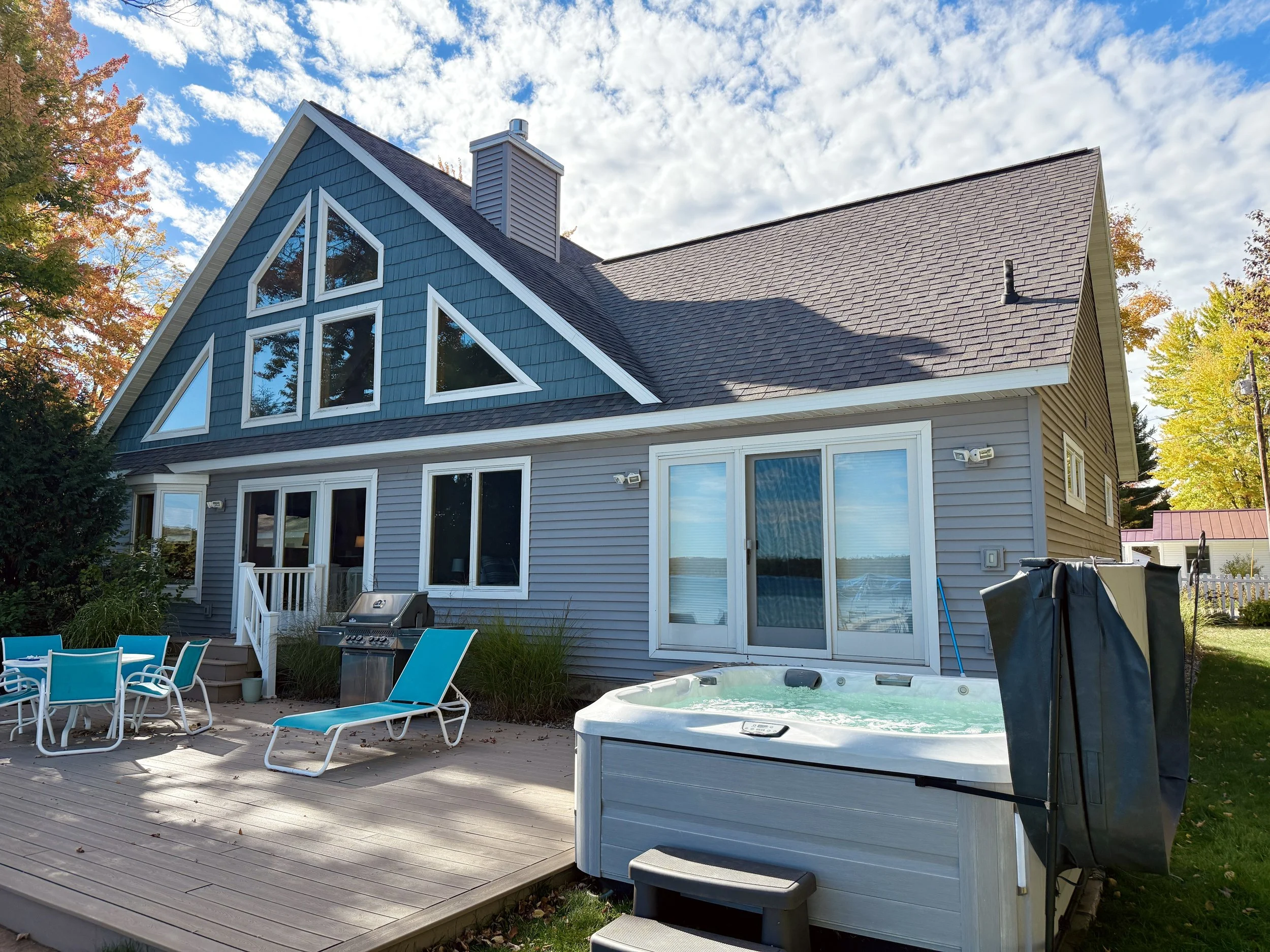 Backyard of a house with a wooden deck, outdoor chairs, a gas grill, and a hot tub. The house has gray siding, large windows, and a roof with dormer windows. There are trees with autumn leaves in the background and a partly cloudy sky.