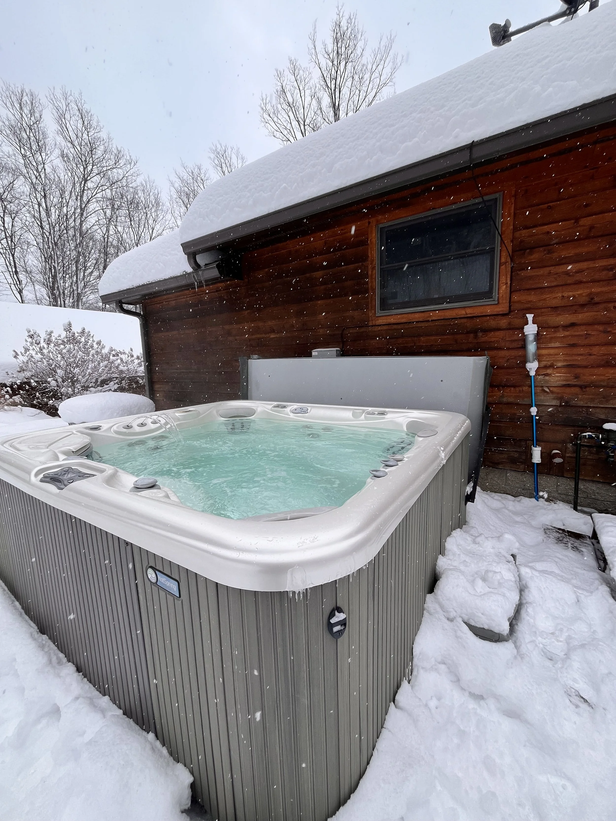 An outdoor hot tub filled with water, surrounded by snow, next to a wooden house with a snow-covered roof and a small window.