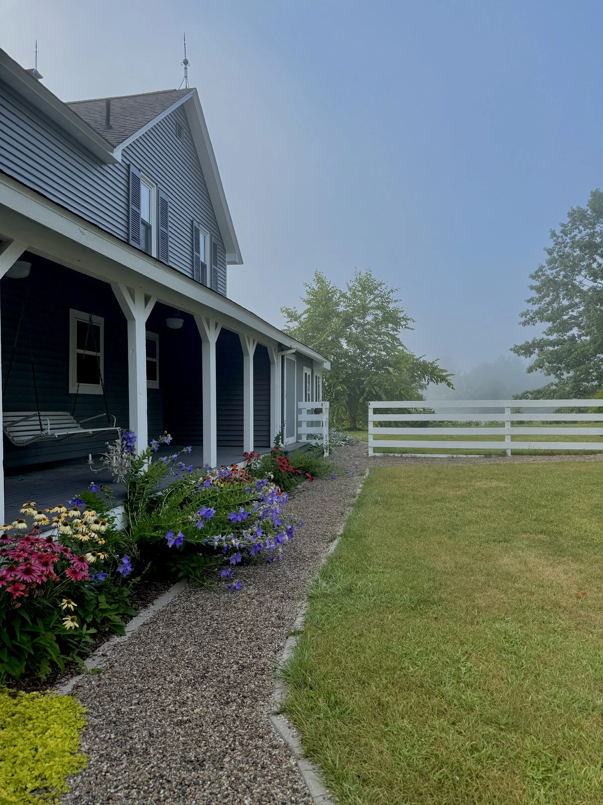 A blue house with a porch, swing, and colorful flower garden, surrounded by a white fence and trees, on a foggy morning.