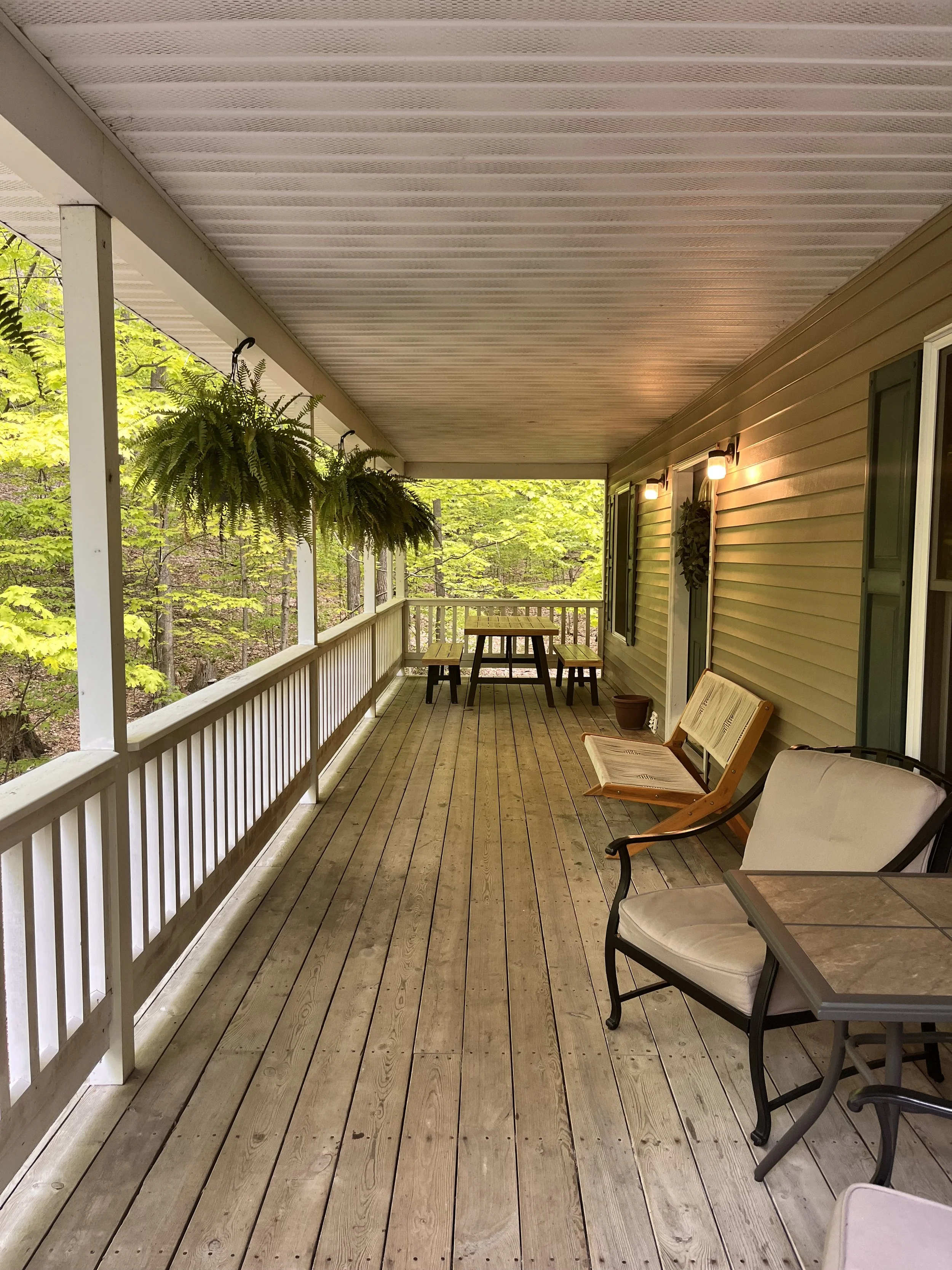 A spacious wooden porch attached to a house with outdoor furniture, hanging ferns, and surrounding trees.