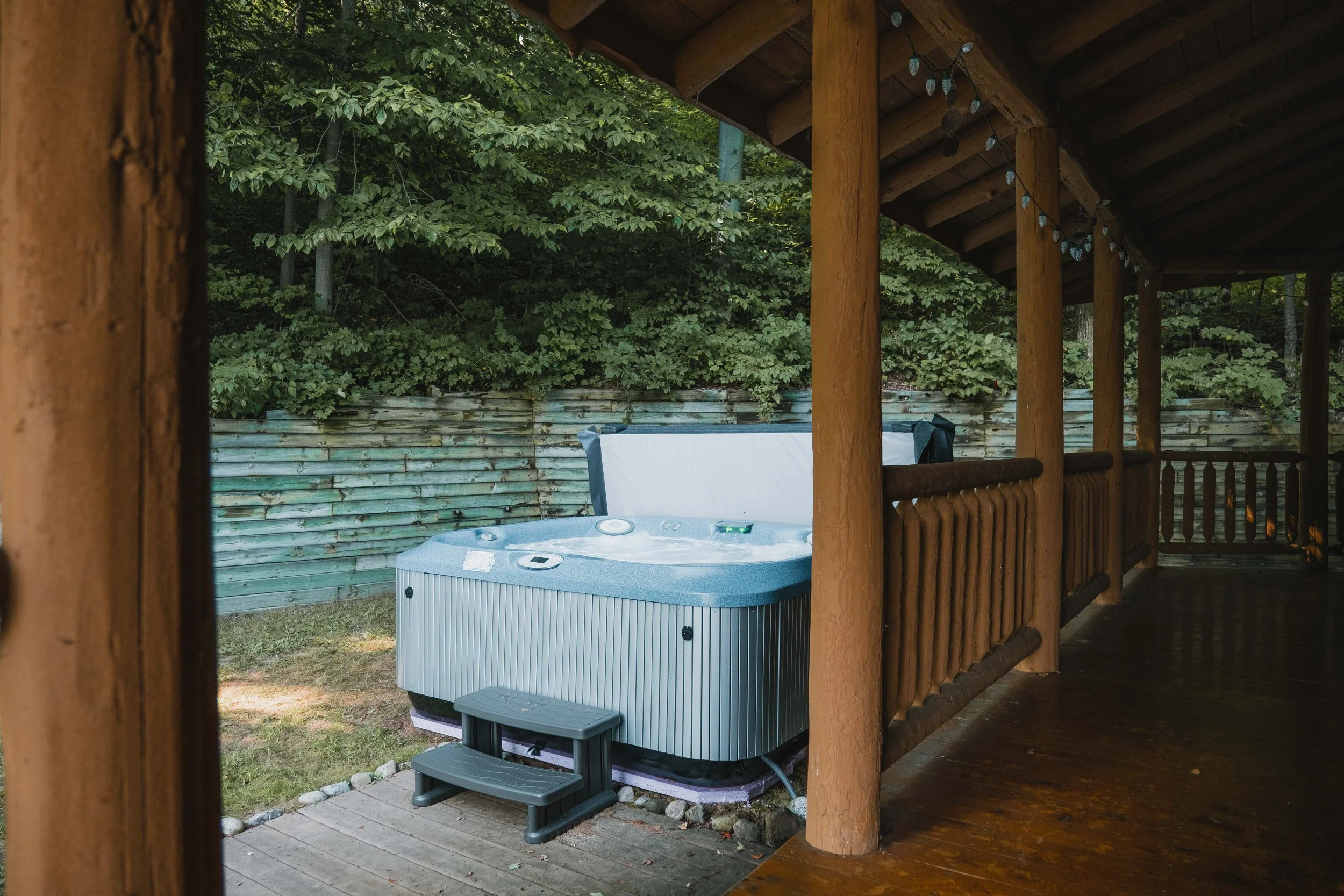Hot tub on a deck outside of a wooden cabin, surrounded by greenery and a wooden fence.