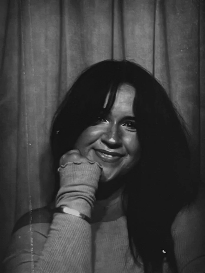 A creative designer woman with long dark hair smiling and resting her cheek on her hand, standing in front of a wooden wall. She offers social media content creation, brand design, web design, and other creative services.