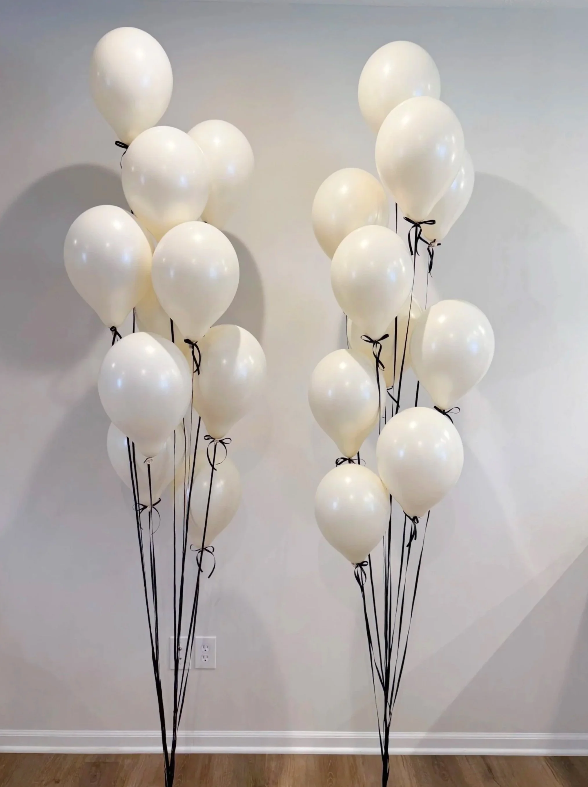 Two clusters of white balloons tied with black ribbons and strings, set against a plain white wall in a room with wooden flooring.