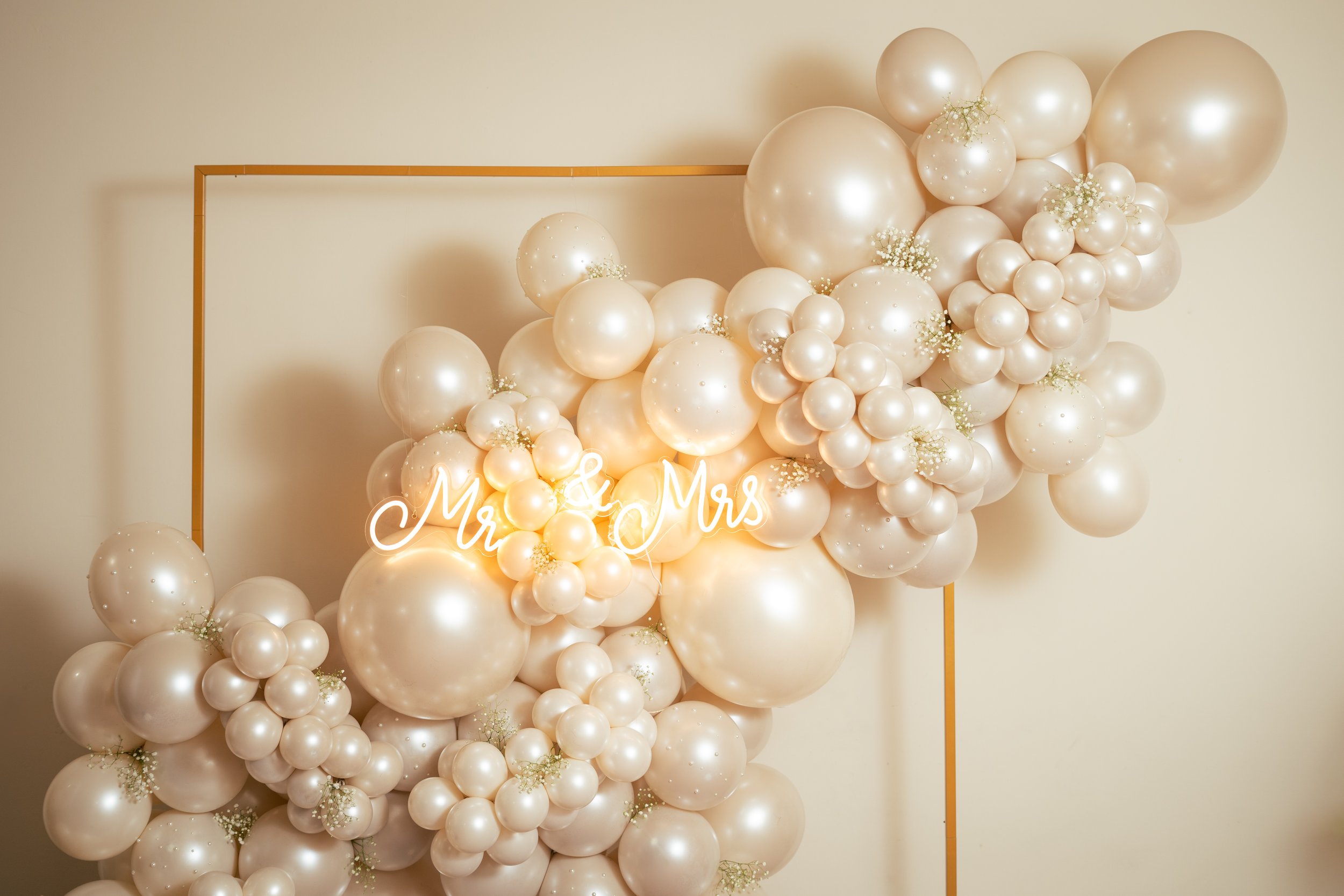 Decorative setup with cream-colored balloons of various sizes, arranged behind a golden rectangular frame. Neon sign saying 'Mr. & Mrs' in cursive font is placed among the balloons, creating an elegant backdrop for a wedding or celebration.