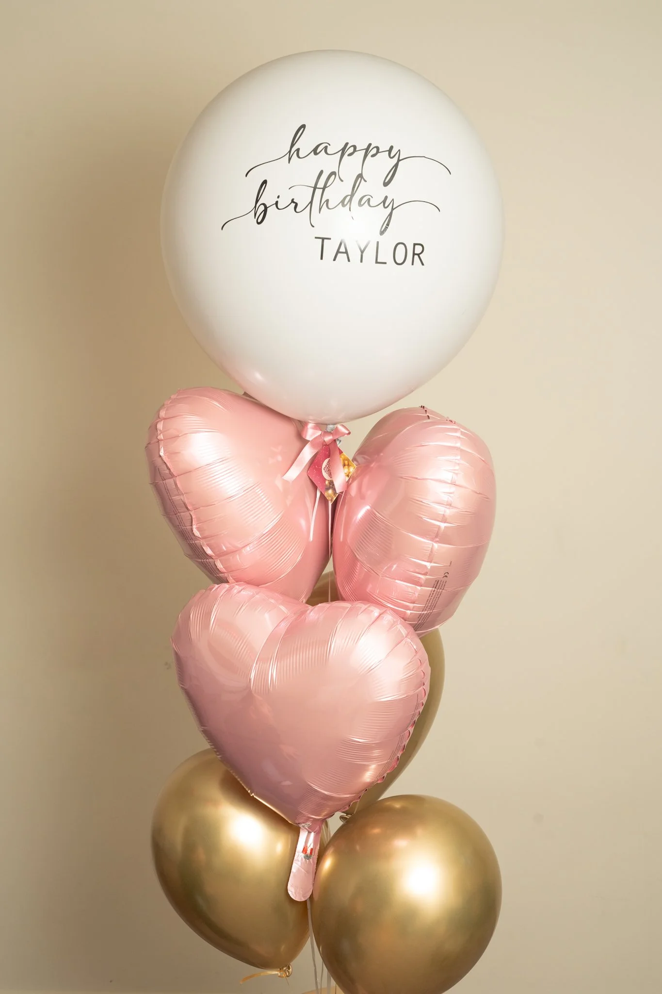 A bouquet of balloons with a large white balloon on top that reads 'happy birthday TAYLOR.' Below it are four pink heart-shaped balloons, and at the bottom are two gold round balloons.