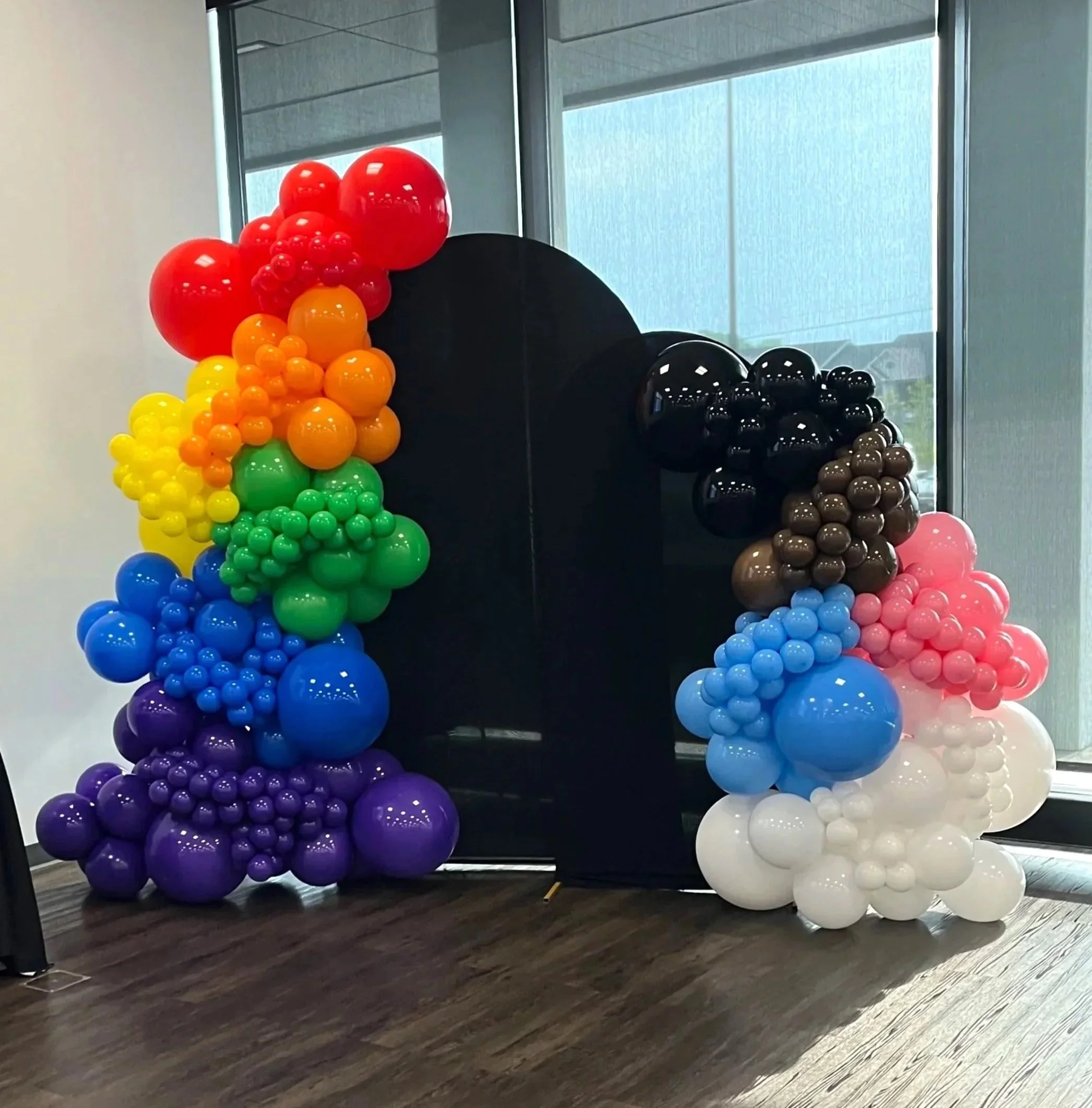 Colorful balloon arch with rainbow colors on the left, black and brown balloons on the right, and a black backdrop in the middle, set up indoors near windows.