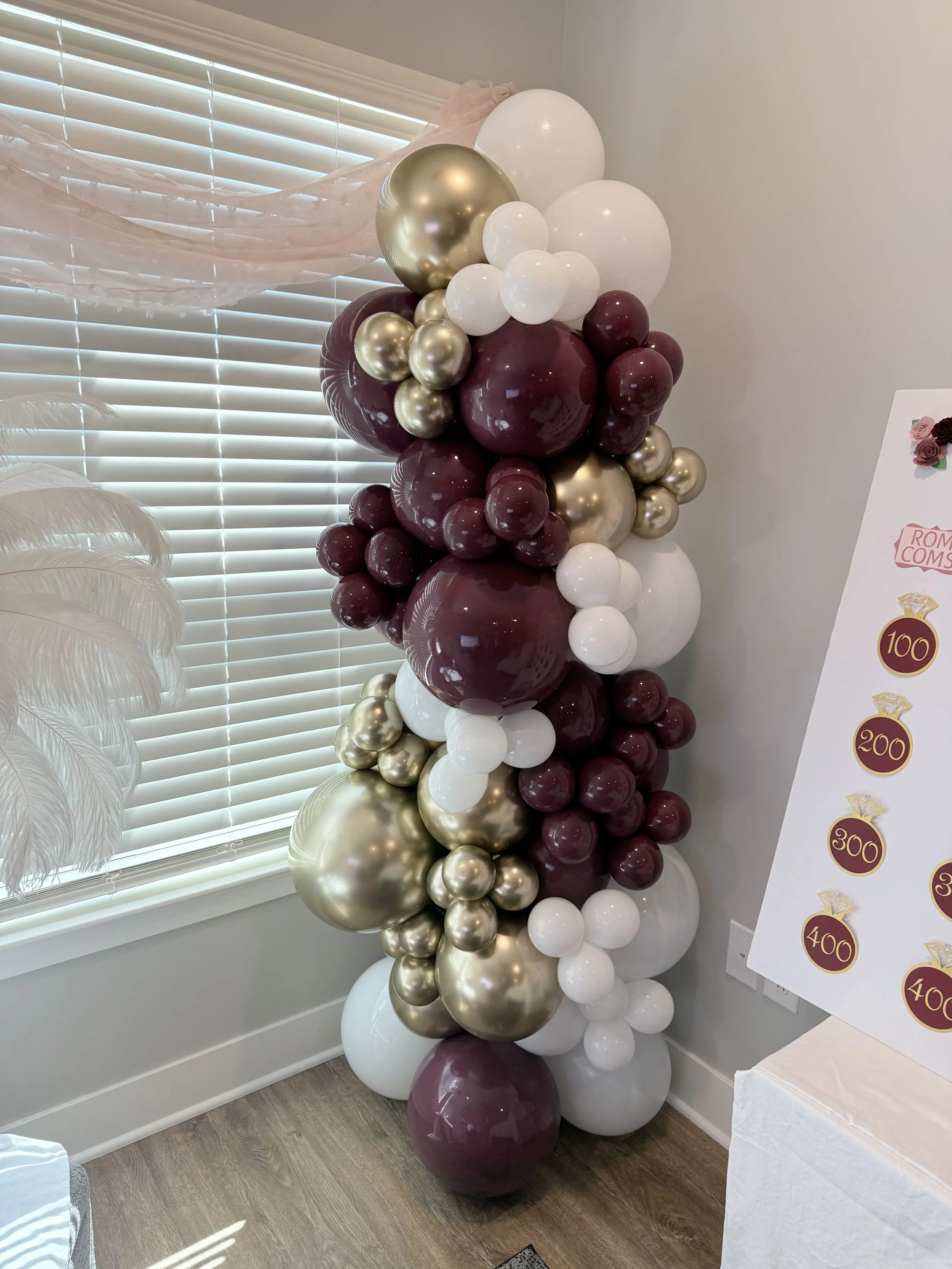 A large decorative balloon arrangement with white, burgundy, and gold balloons, situated near a window with blinds. The arrangement is part of a celebratory event setup.