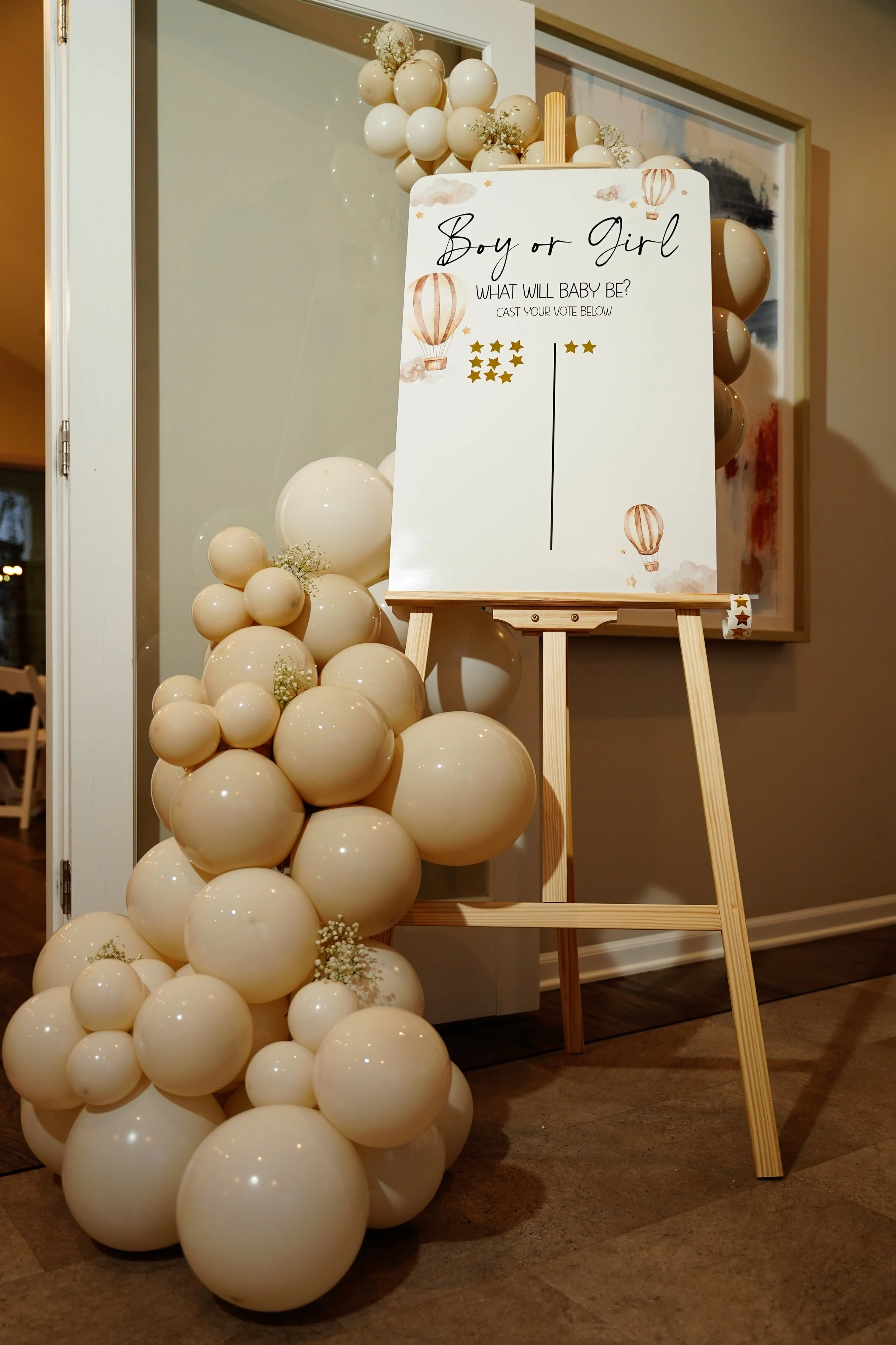 A decorative display for a gender reveal event, featuring a whiteboard on an easel with hot air balloon illustrations and the words "Boy or Girl, What will baby be? cast your vote below," surrounded by cream-colored balloons and small white flowers.